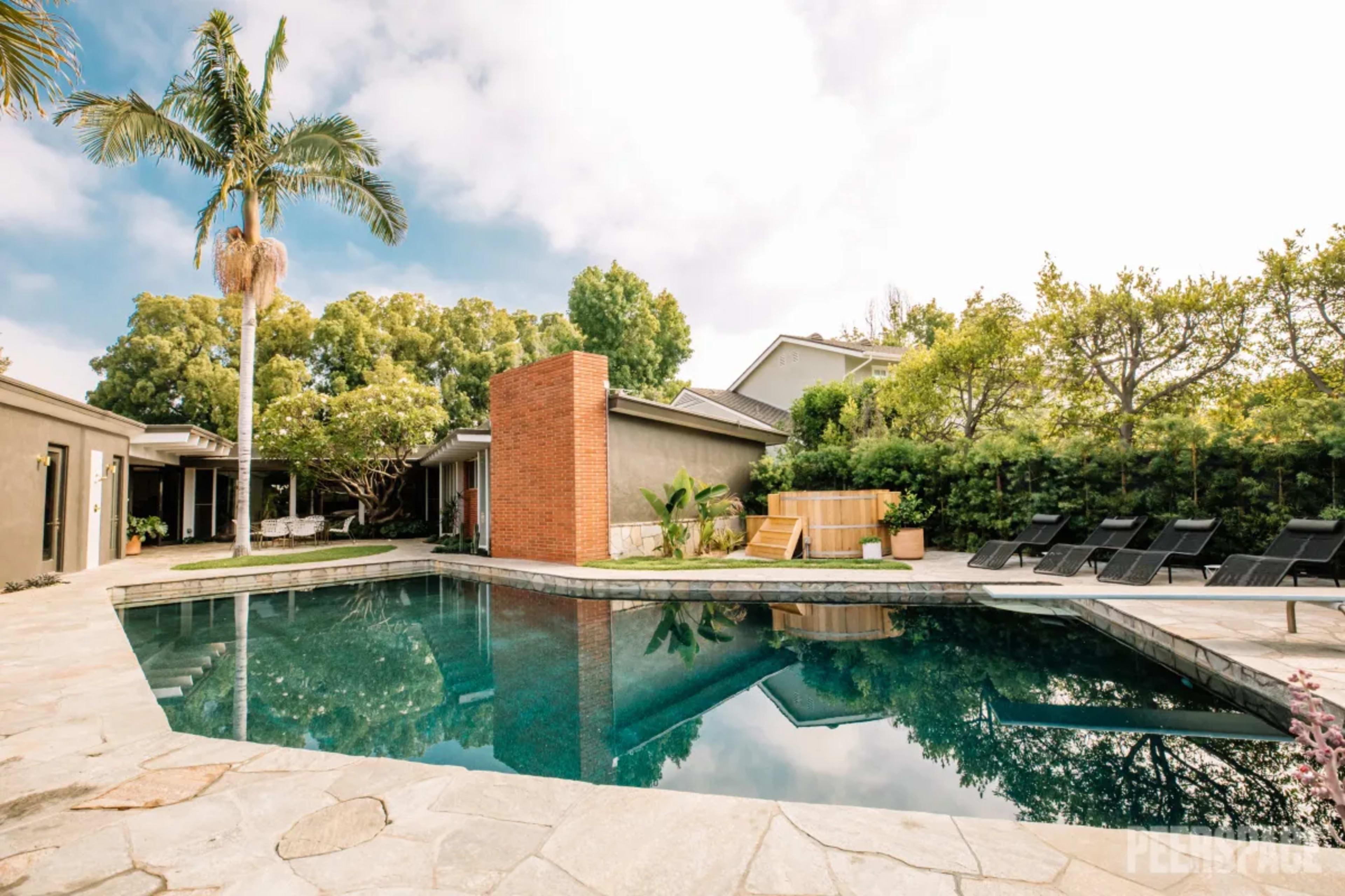 The image shows a modern outdoor pool surrounded by a stone patio, with a brick feature wall and lounge chairs nearby, set among lush greenery.