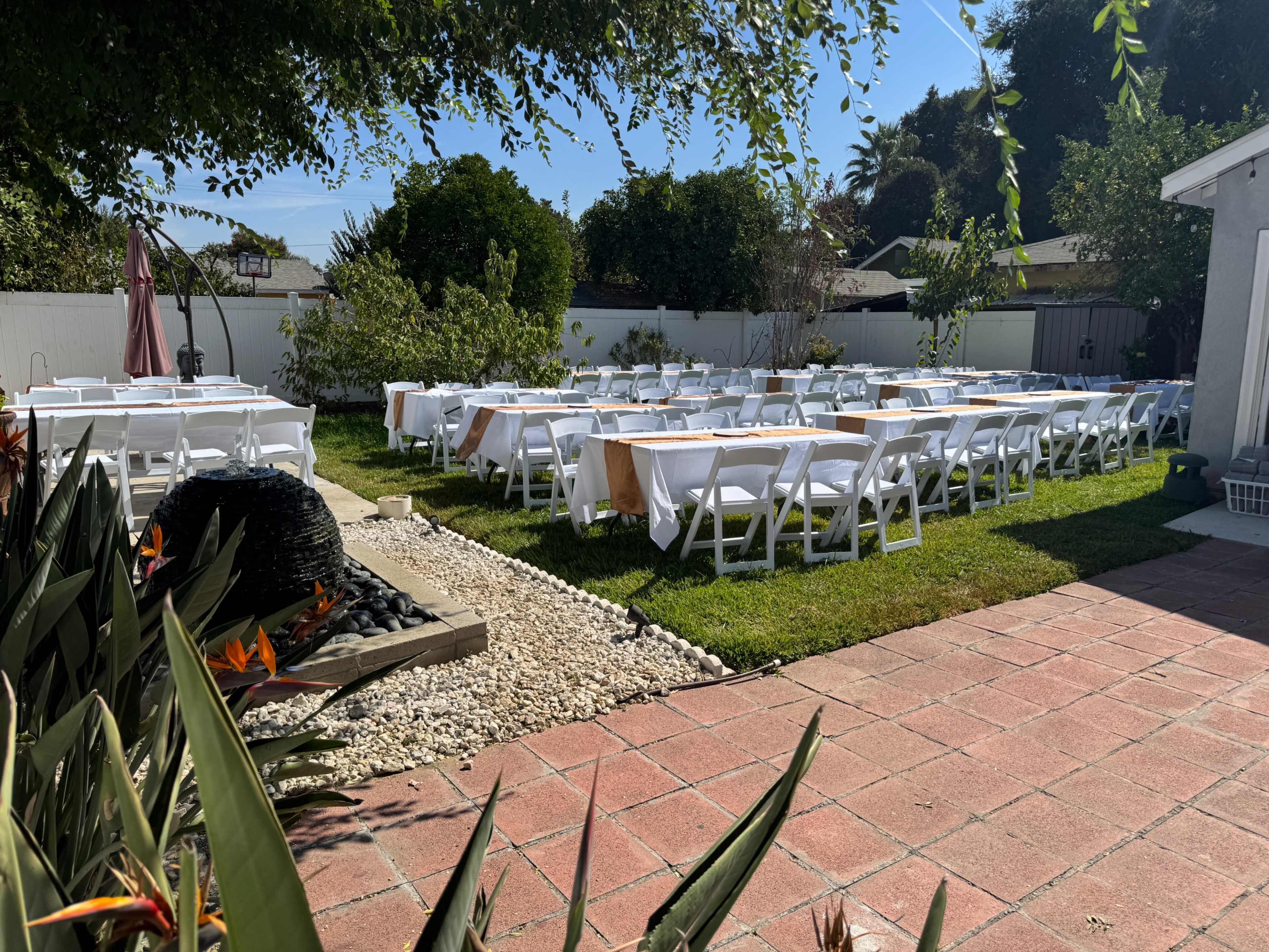 A garden setting features multiple rows of white tables and chairs arranged on grass, surrounded by plants and a stone path.