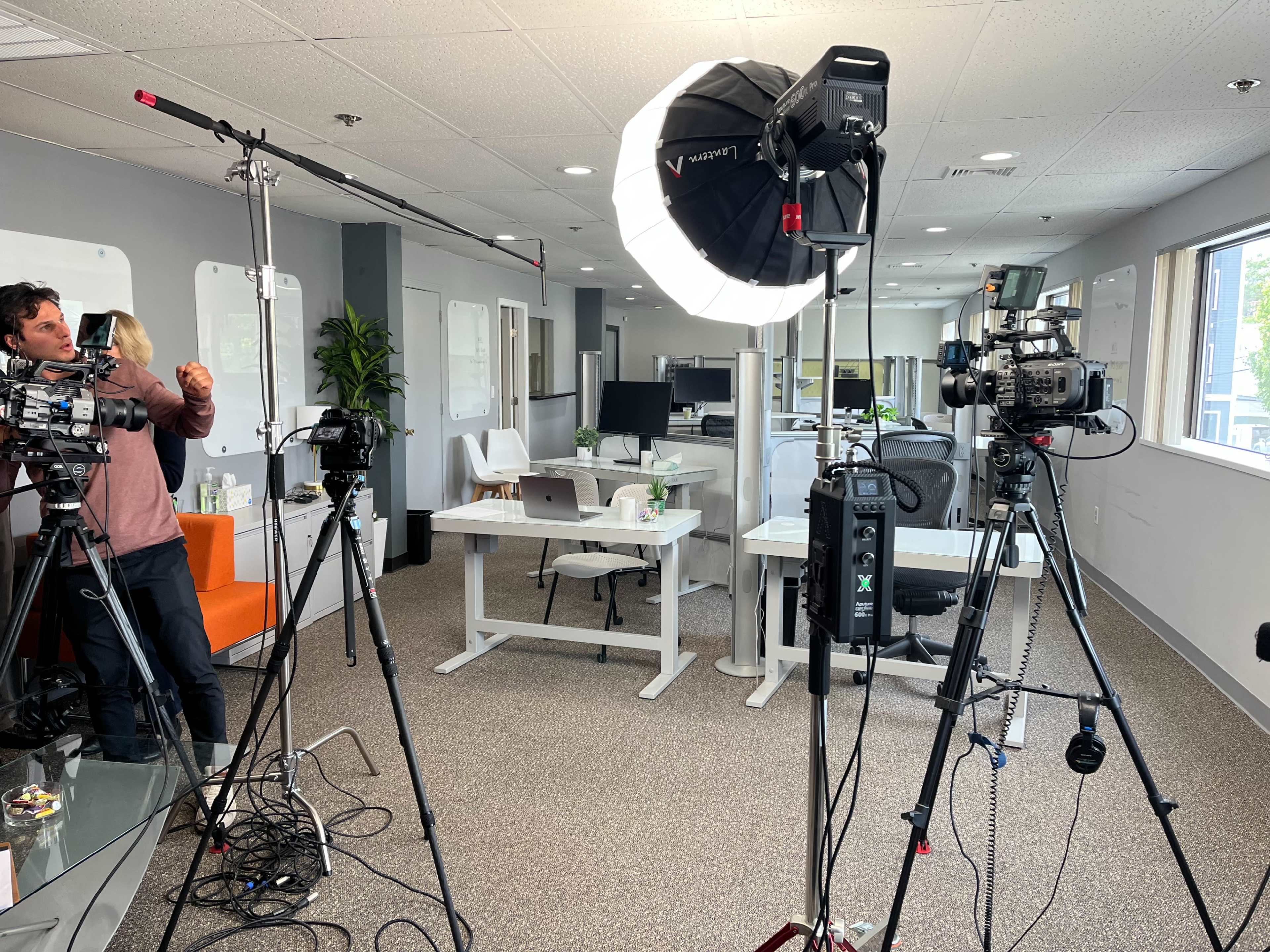 The image shows a modern office space equipped with multiple cameras and lighting setups for a filming project, with crew members adjusting the equipment.