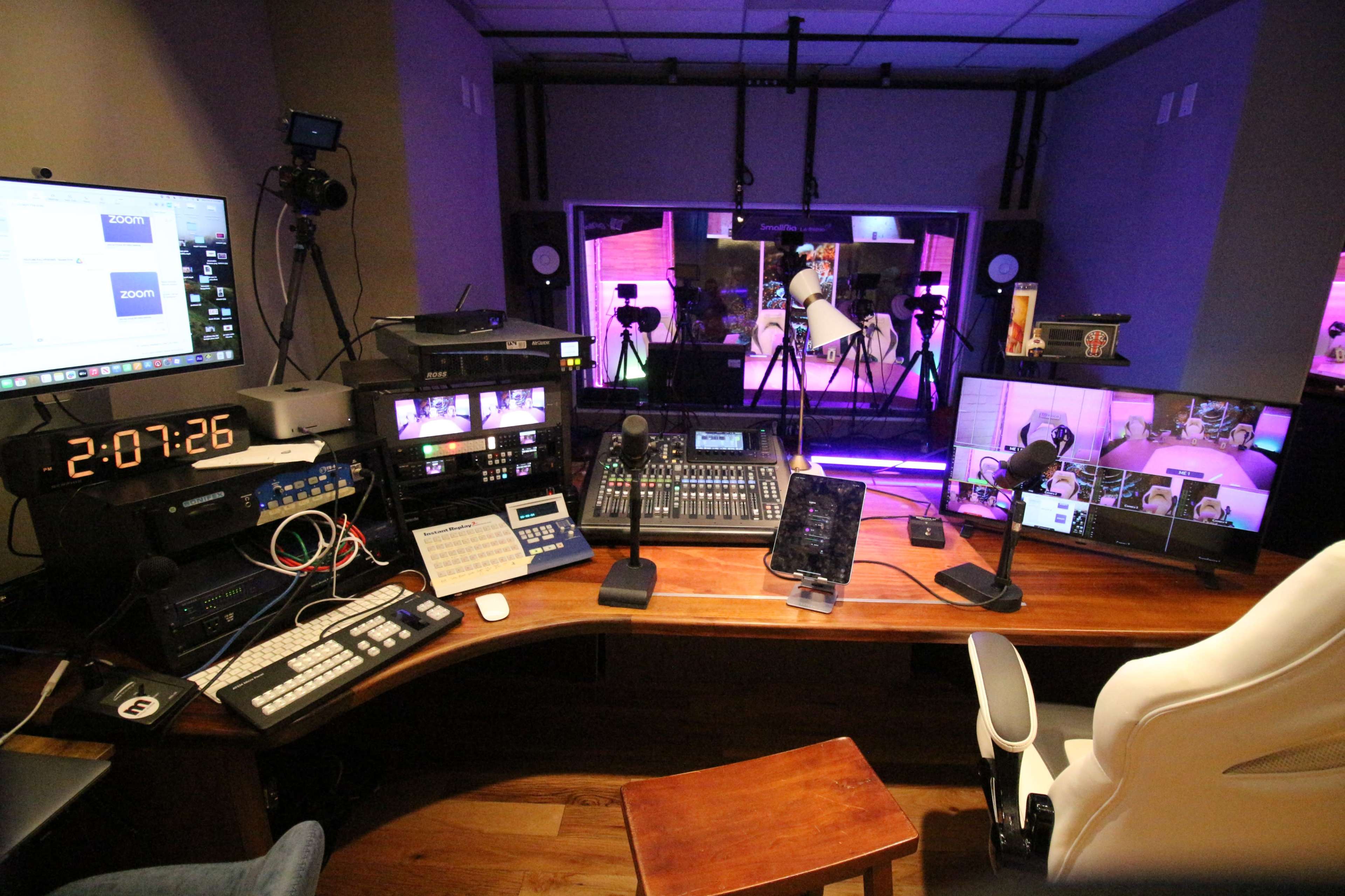 The image shows a broadcast control room equipped with multiple monitors, audio equipment, and a camera setup in a dimly lit environment.