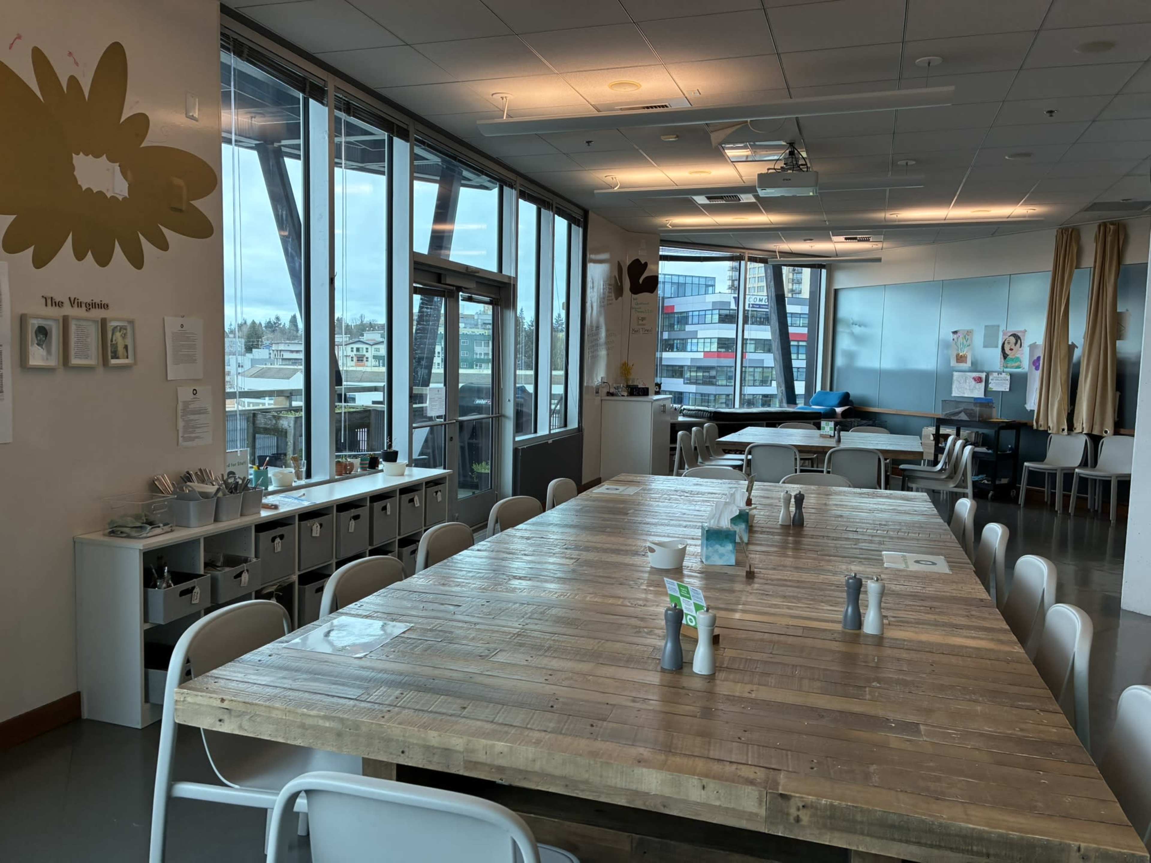 A large wooden table surrounded by white chairs is set in a bright room with large windows, showing a view of buildings outside.