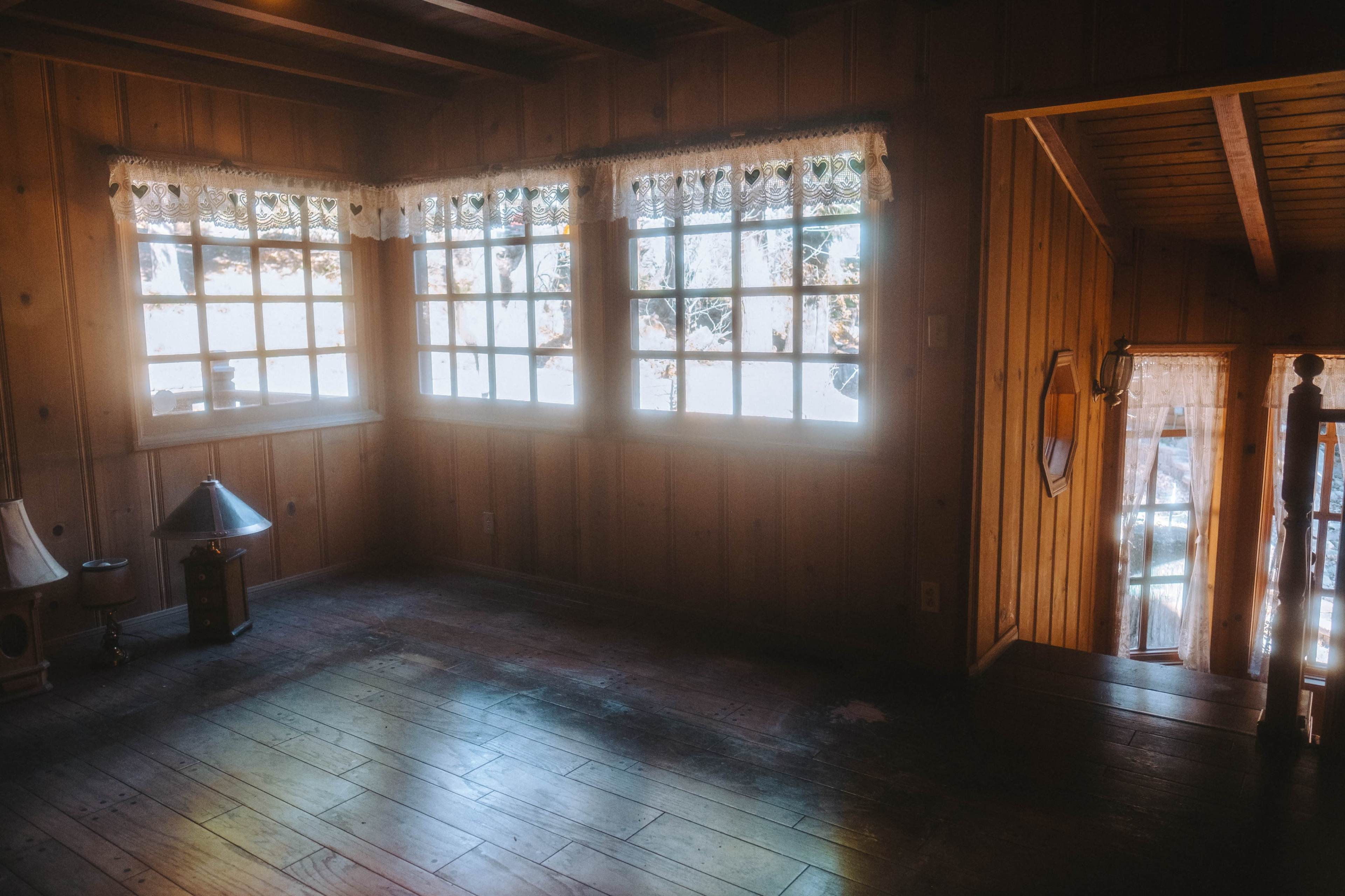 A wood-paneled room features multiple windows with lace curtains, illuminating the space with natural light.