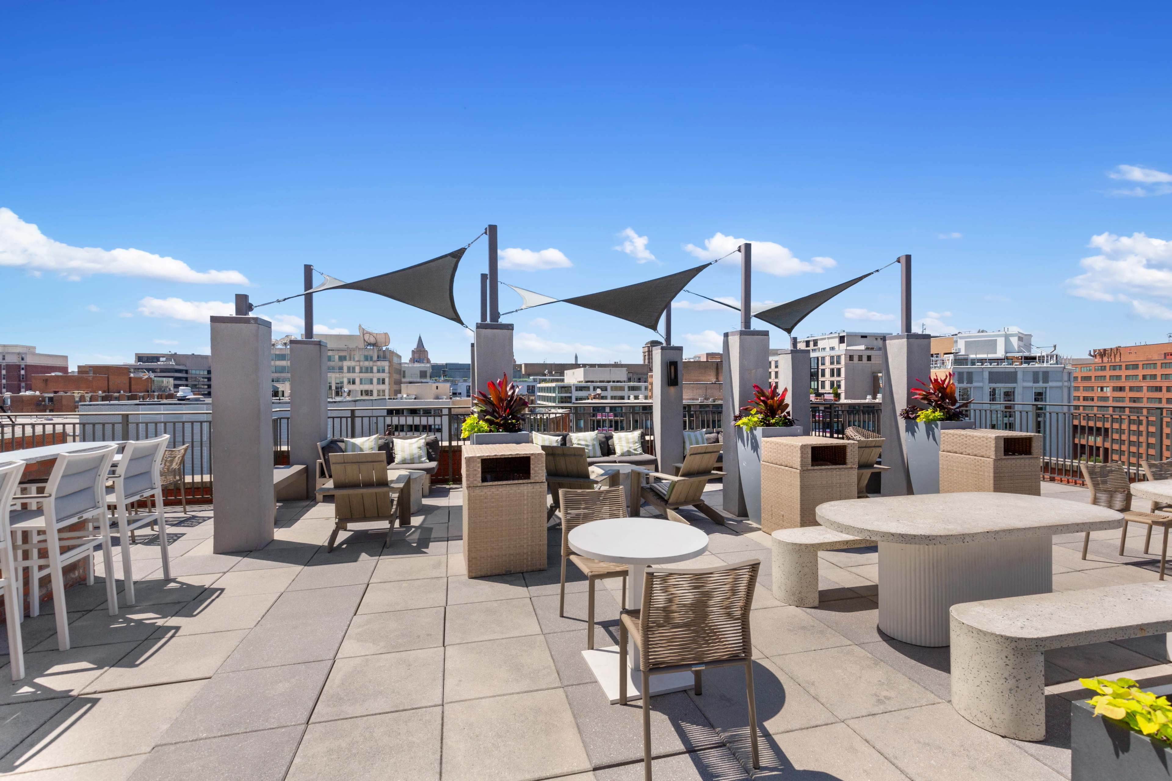 The image shows a rooftop patio area with modern furniture, including tables and chairs, along with decorative planters and shade sails overhead.