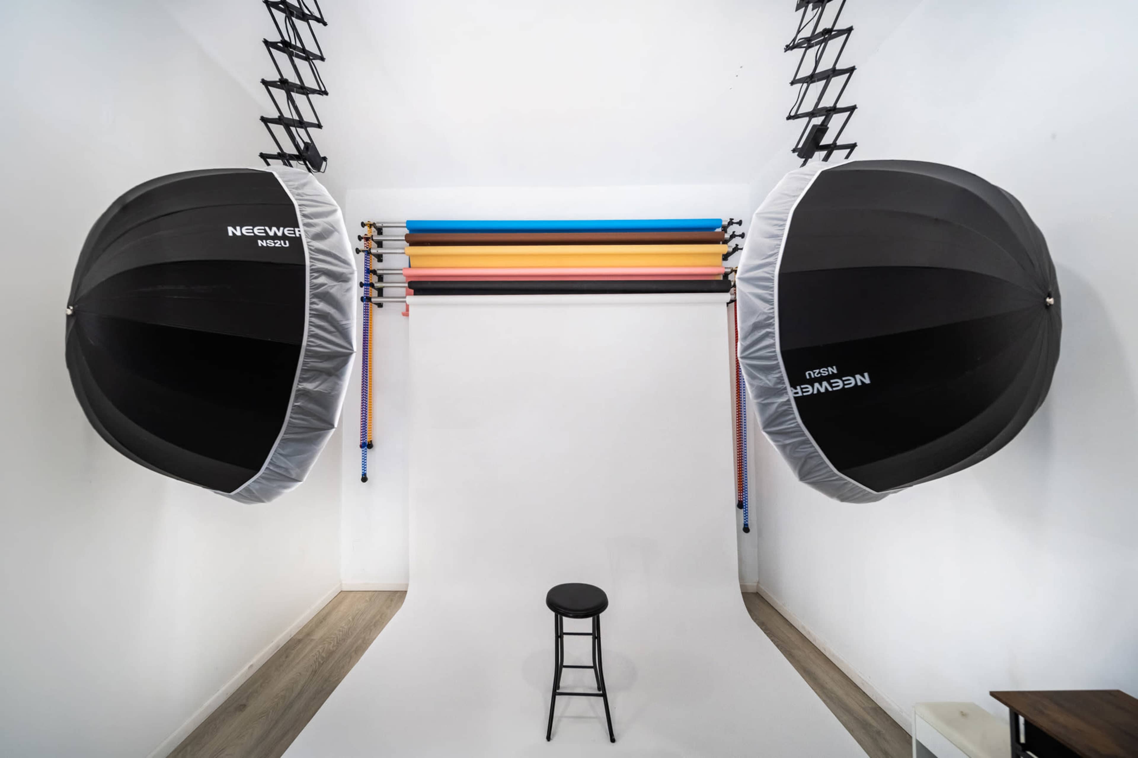 The image shows a minimalist photography studio featuring a white backdrop, two large softbox lights, and a single black stool in the center.