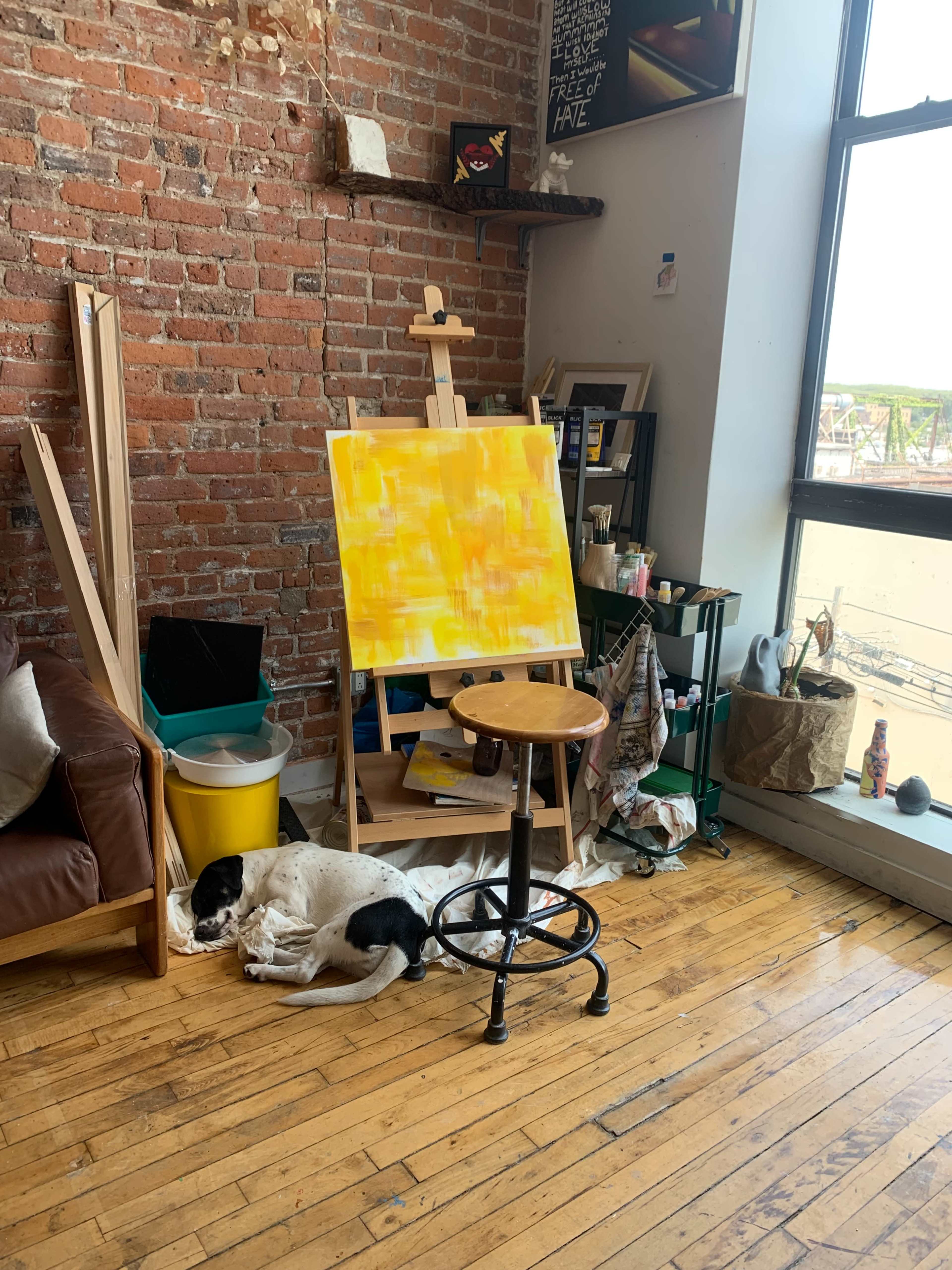 A dog lies curled up on the wooden floor beside an easel with an unfinished yellow painting in a cozy art studio.
