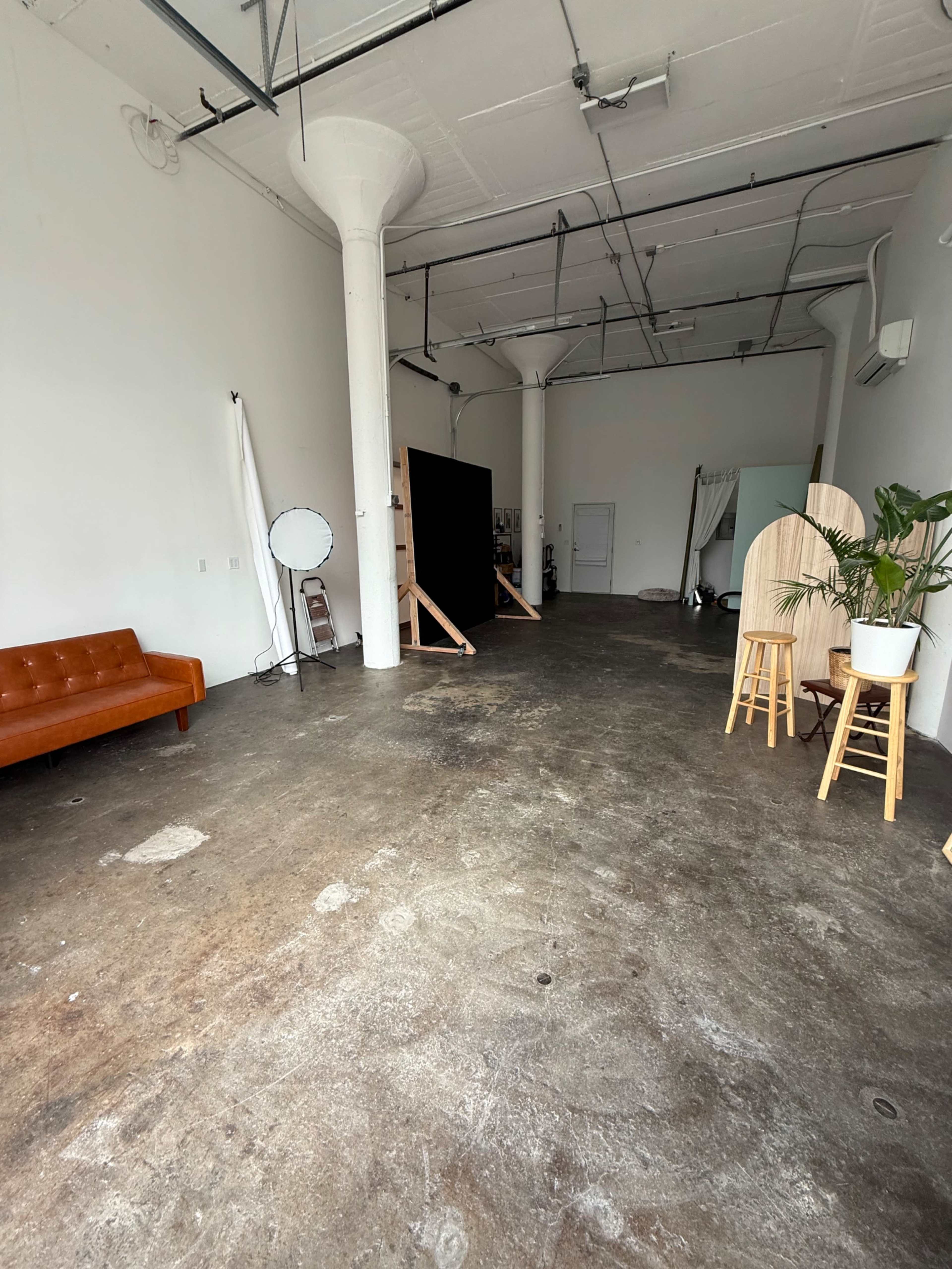 The image shows a spacious, minimally furnished interior with a brown sofa, two wooden stools, a large black backdrop, and a potted plant.