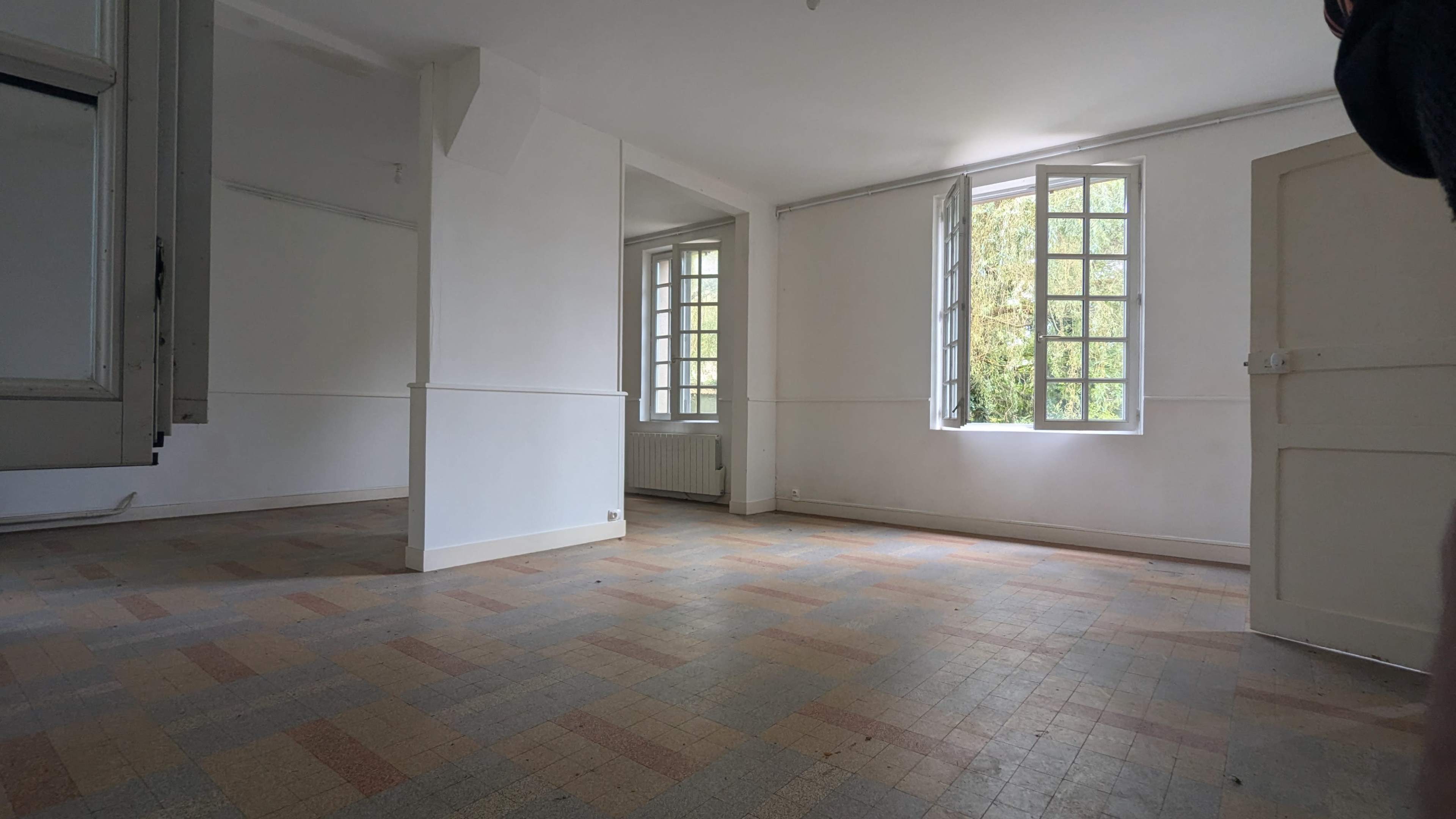 The image shows an empty room with large windows, light-colored walls, and a patterned floor.