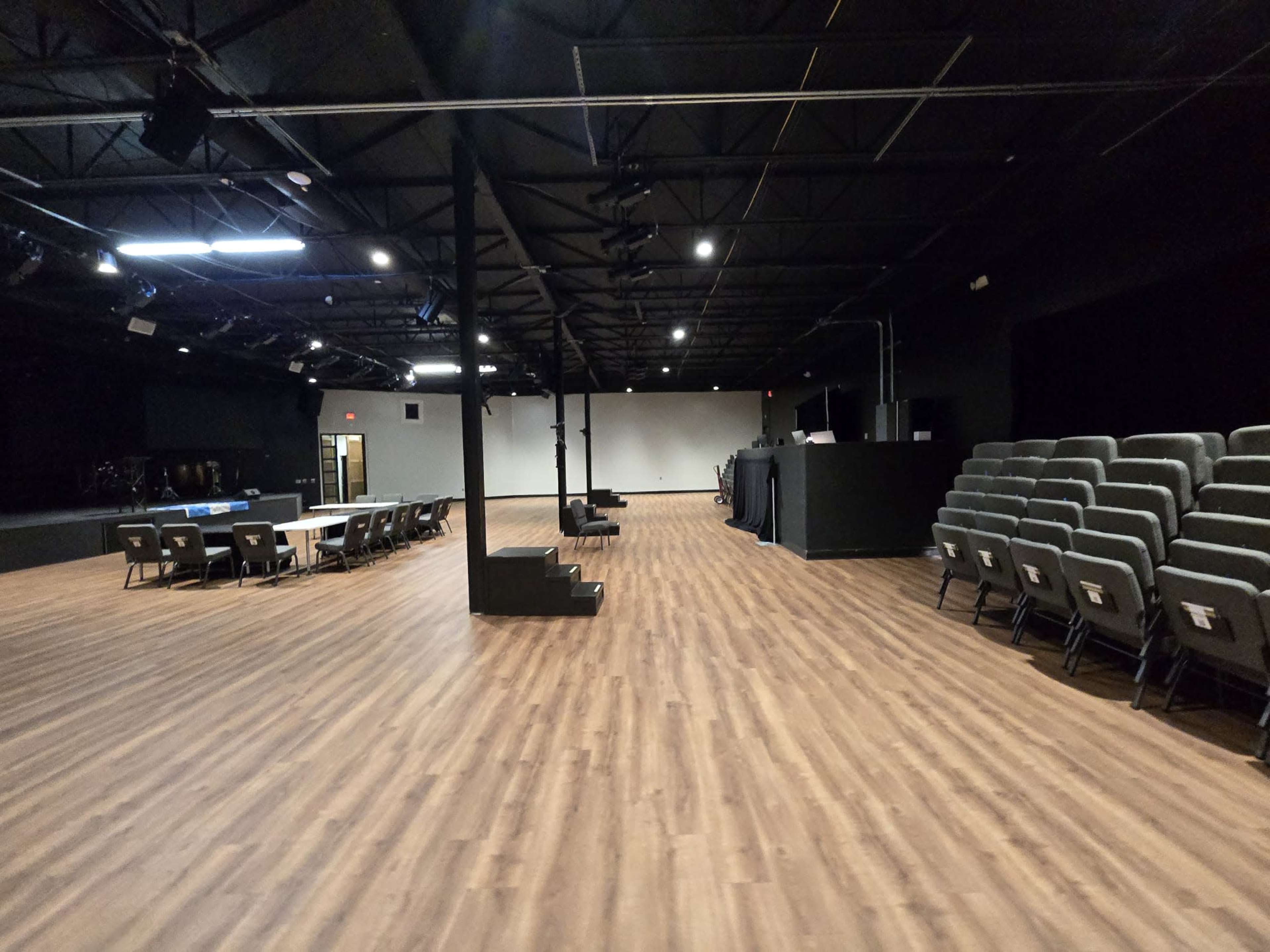 The image shows a spacious, empty auditorium with wooden flooring, rows of chairs, and a stage area at the front.