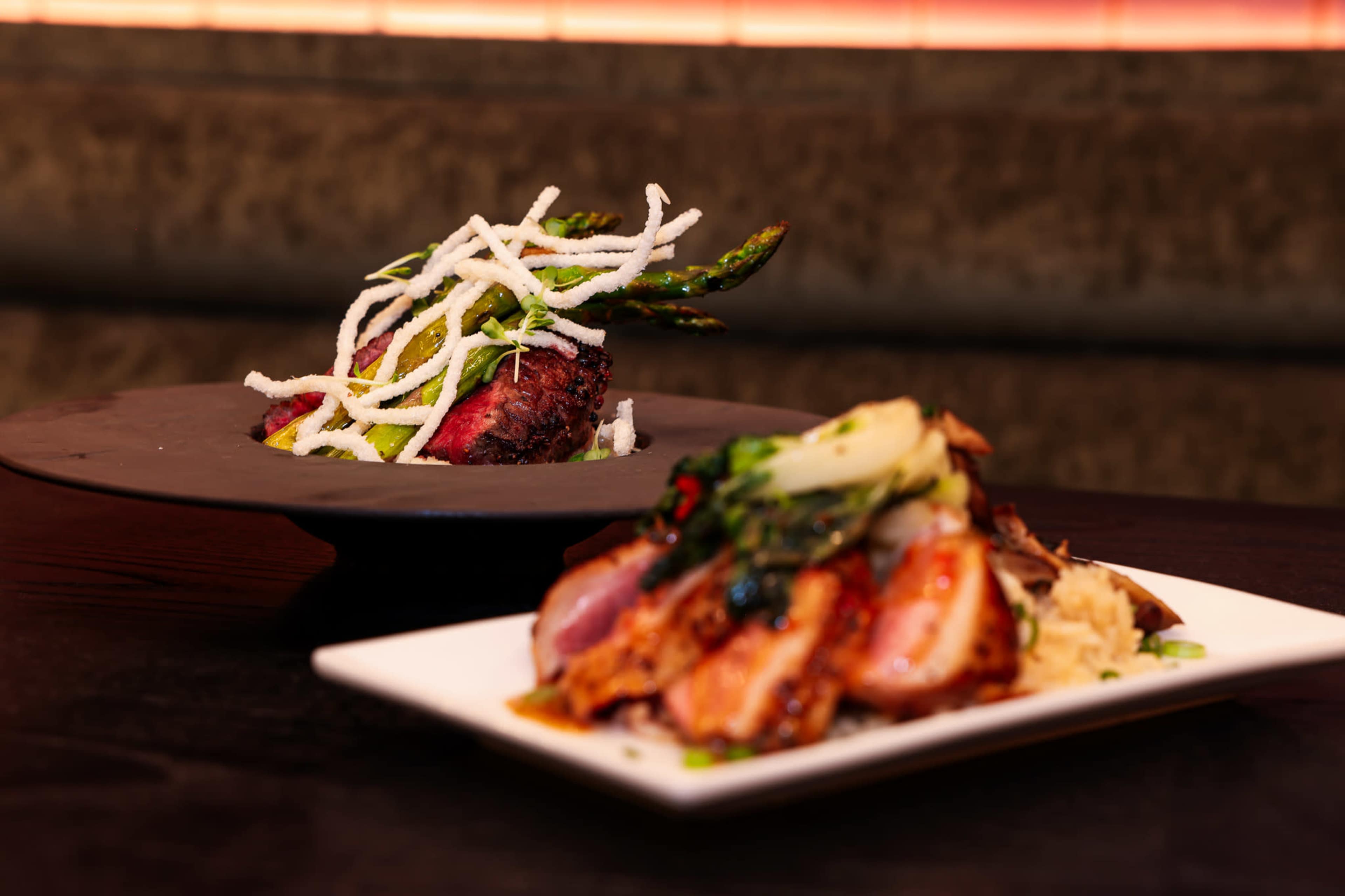 The image features two plated dishes: one with grilled meat topped with asparagus and crispy noodles, and another with sliced meat accompanied by vegetables on a bed of rice.