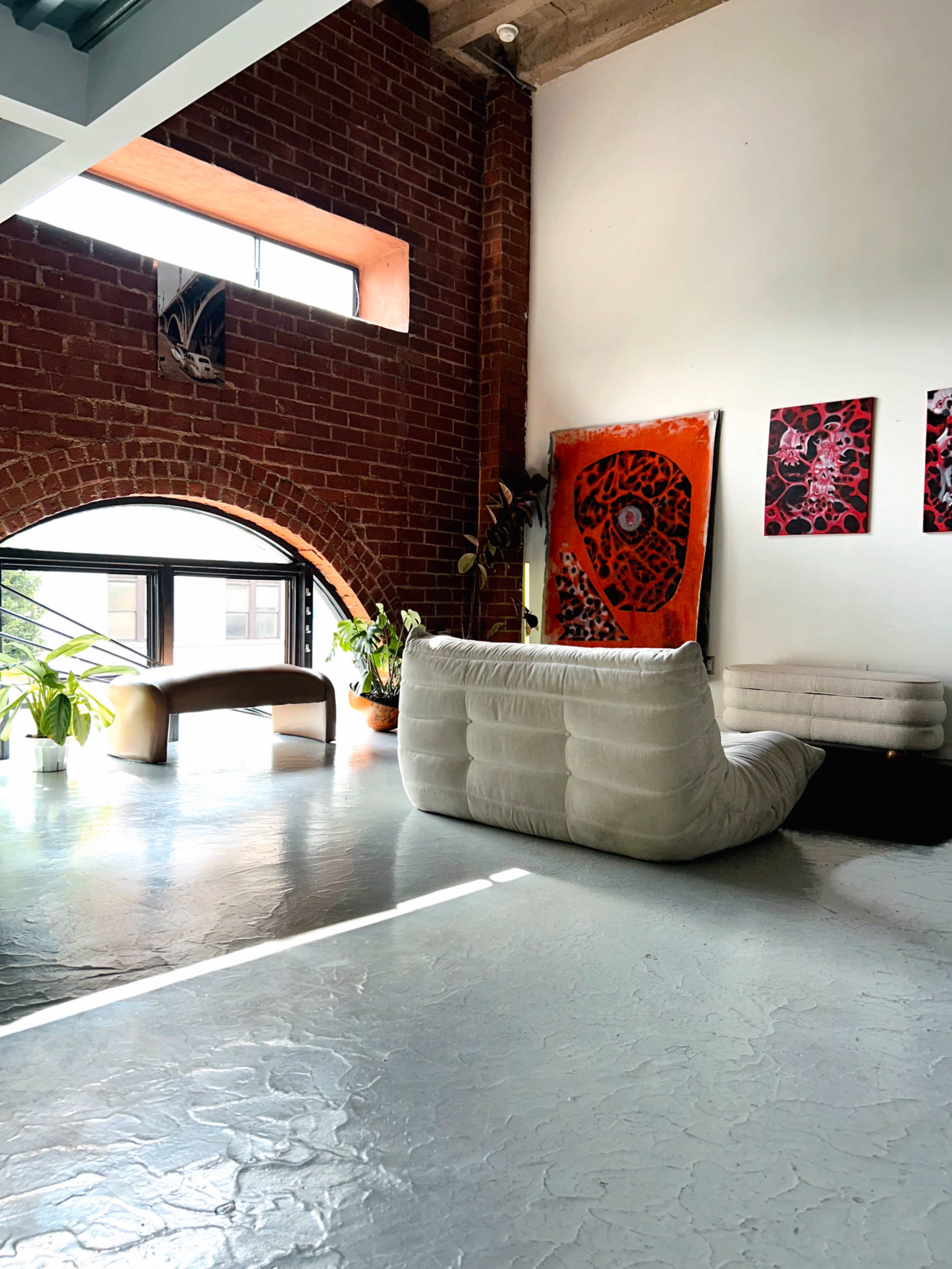 The image shows a spacious room with exposed brick walls, a large window, and modern furniture, including a white couch and artistic wall decor.