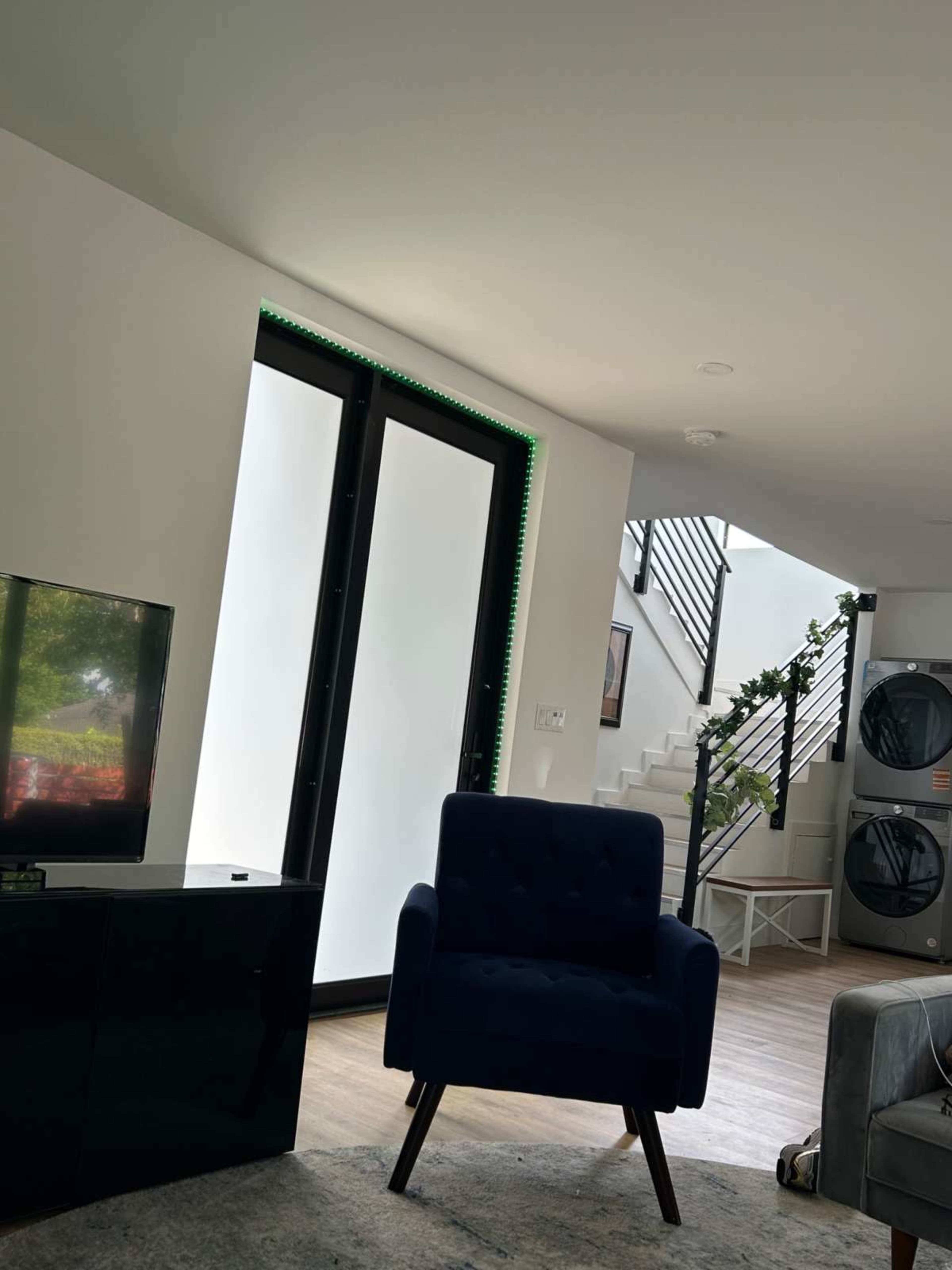 The image shows a modern living space featuring a blue armchair, a television, and a staircase leading to an upper level, with large windows illuminated by green lights.