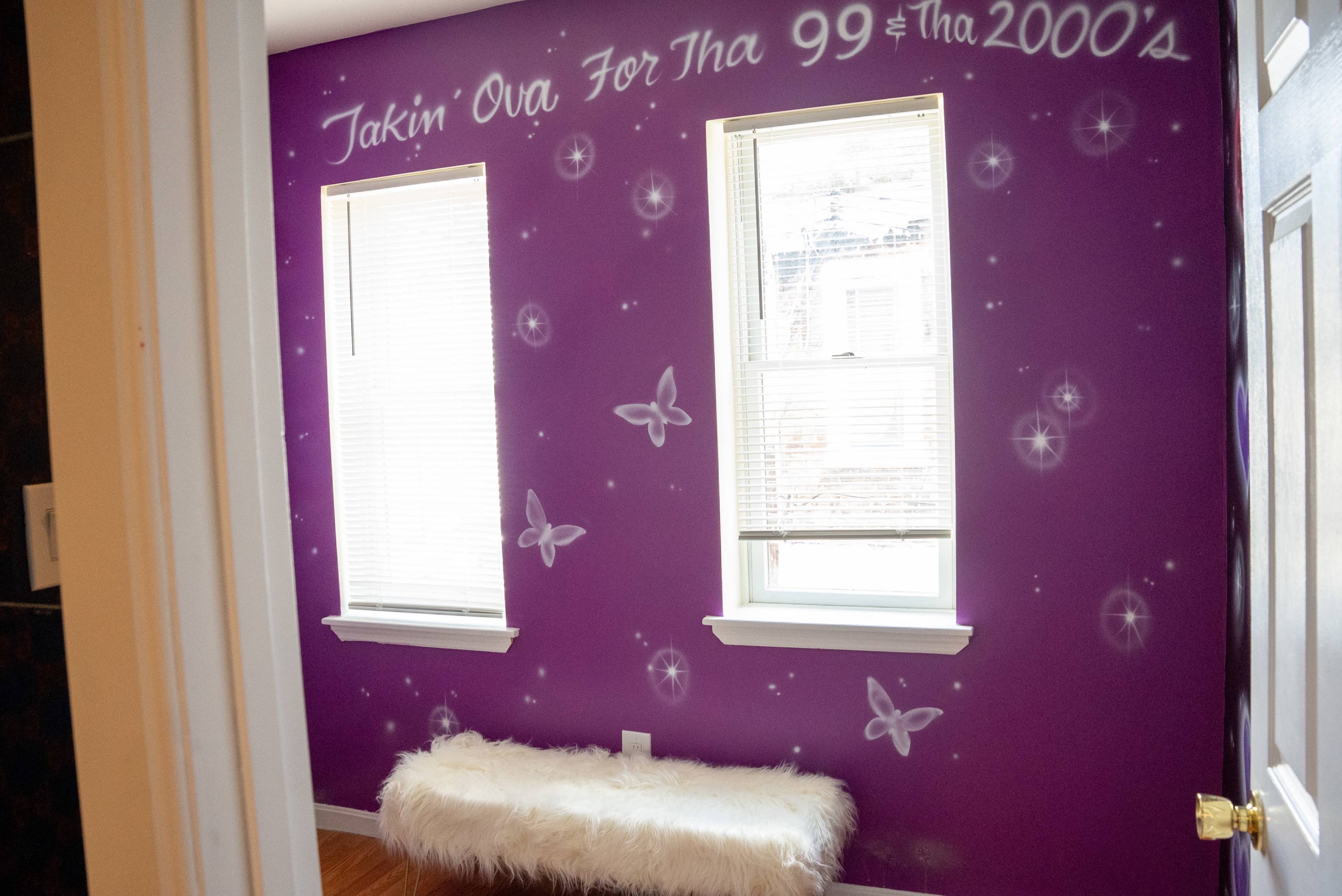 The image shows a small room with purple walls decorated with white butterflies and text, featuring two windows and a white fur bench.