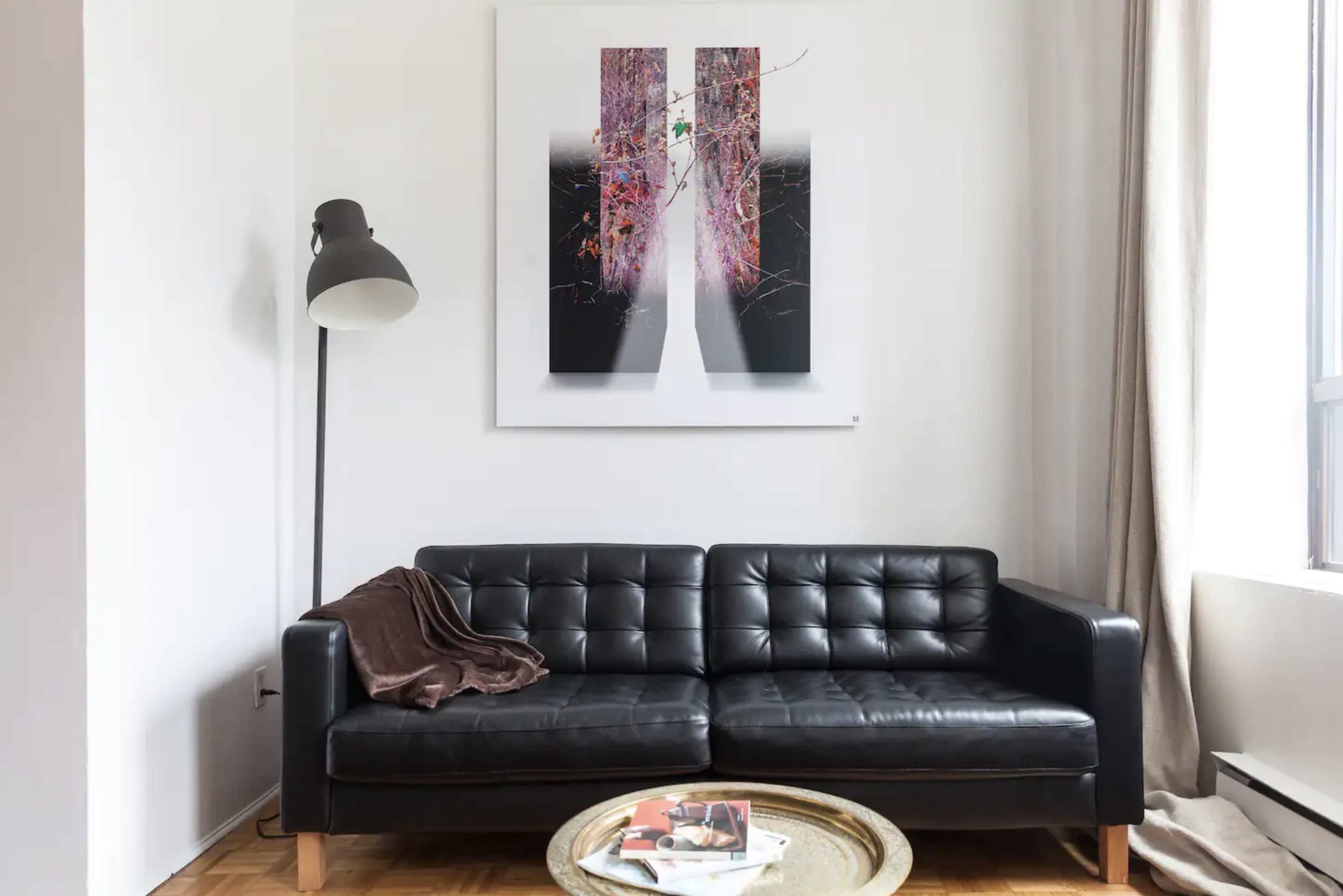 A black leather sofa sits against a white wall, accompanied by a large abstract artwork and a small coffee table with magazines.