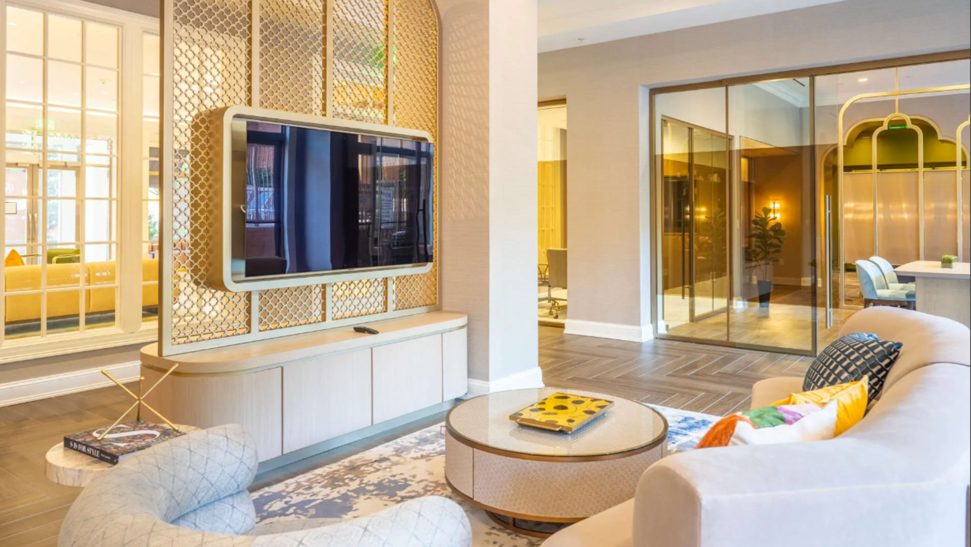 The image shows a modern lounge area featuring a television mounted on a decorative wall unit, with a cozy seating arrangement and glass doors leading to another room.