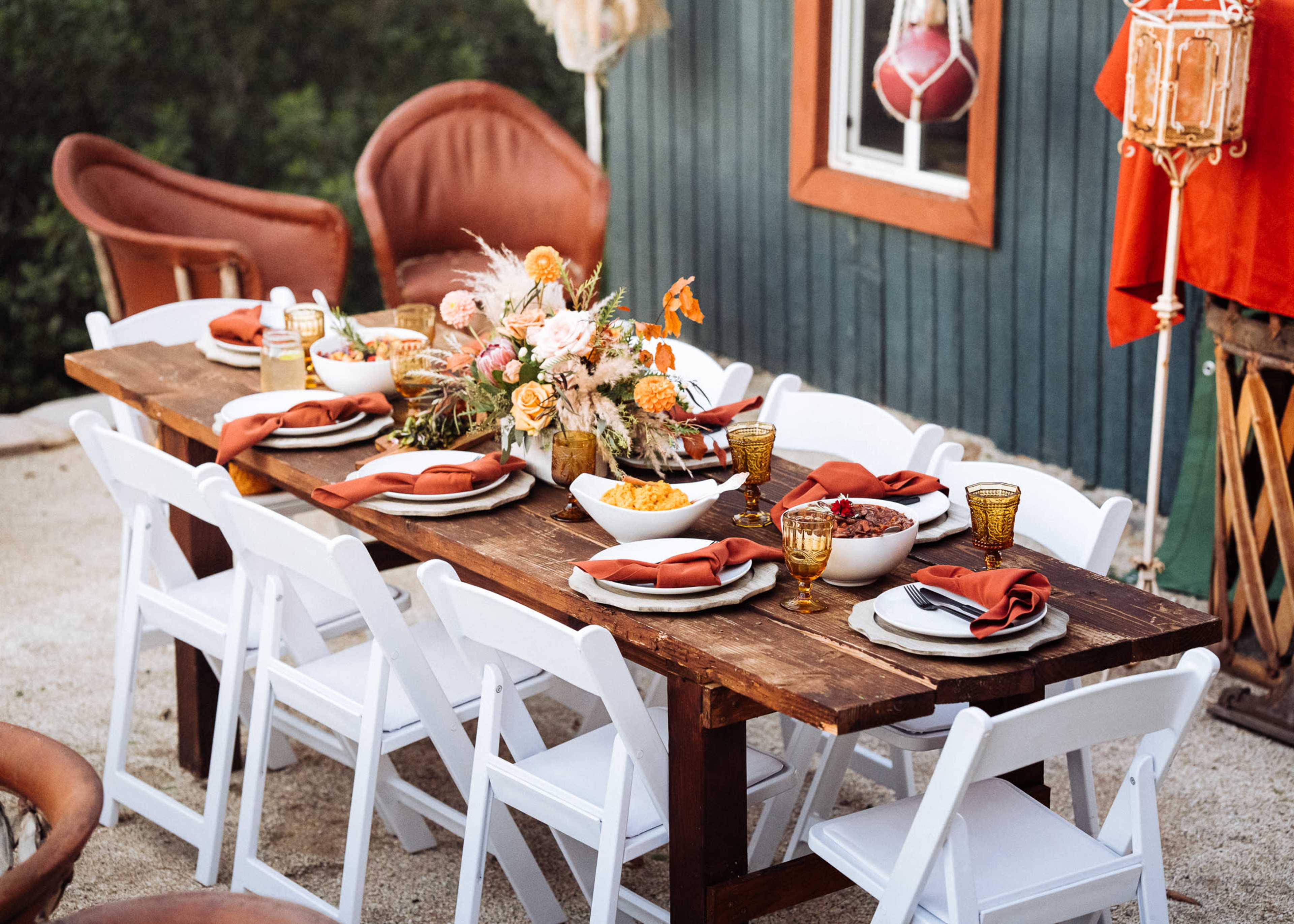 A wooden table is set with plates, glasses, and napkins, surrounded by white chairs, and decorated with a floral centerpiece in an outdoor setting.