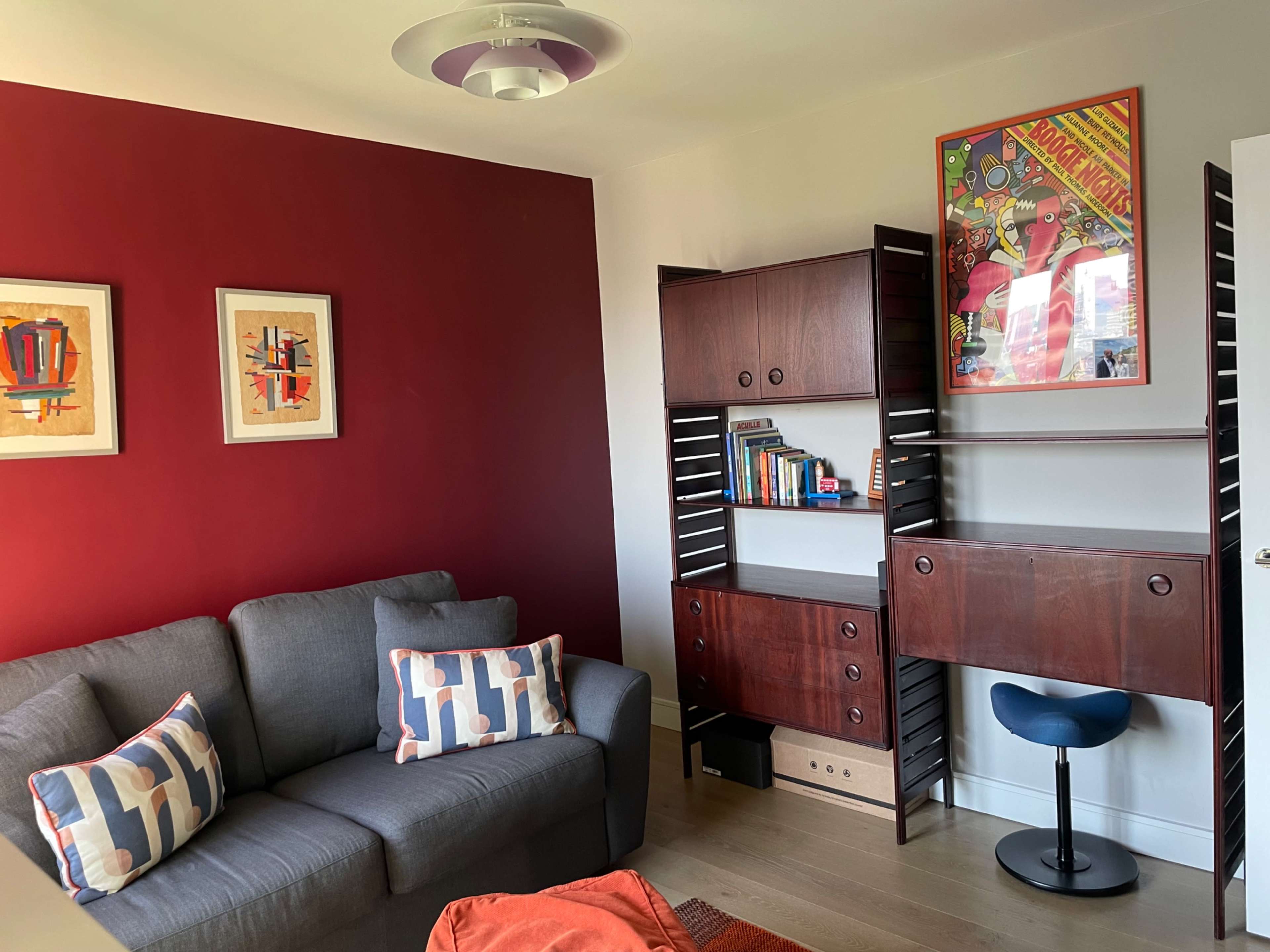 A living room with a gray sofa, two framed artworks on a red wall, and a wooden shelving unit containing books and a small desk.