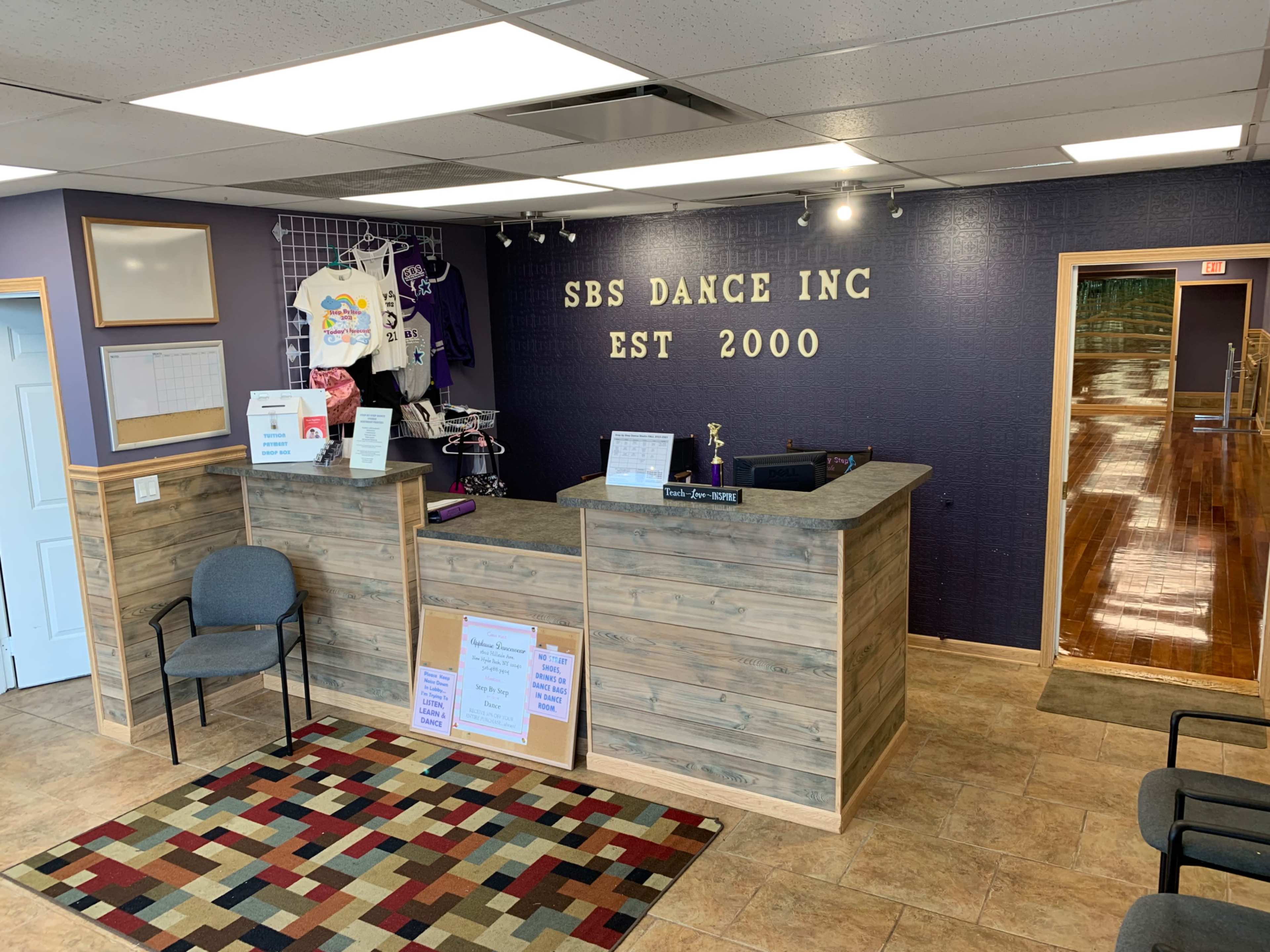 A reception area for SBS Dance Inc., featuring a wooden front desk, promotional items on display, and a seating area with a patterned rug.