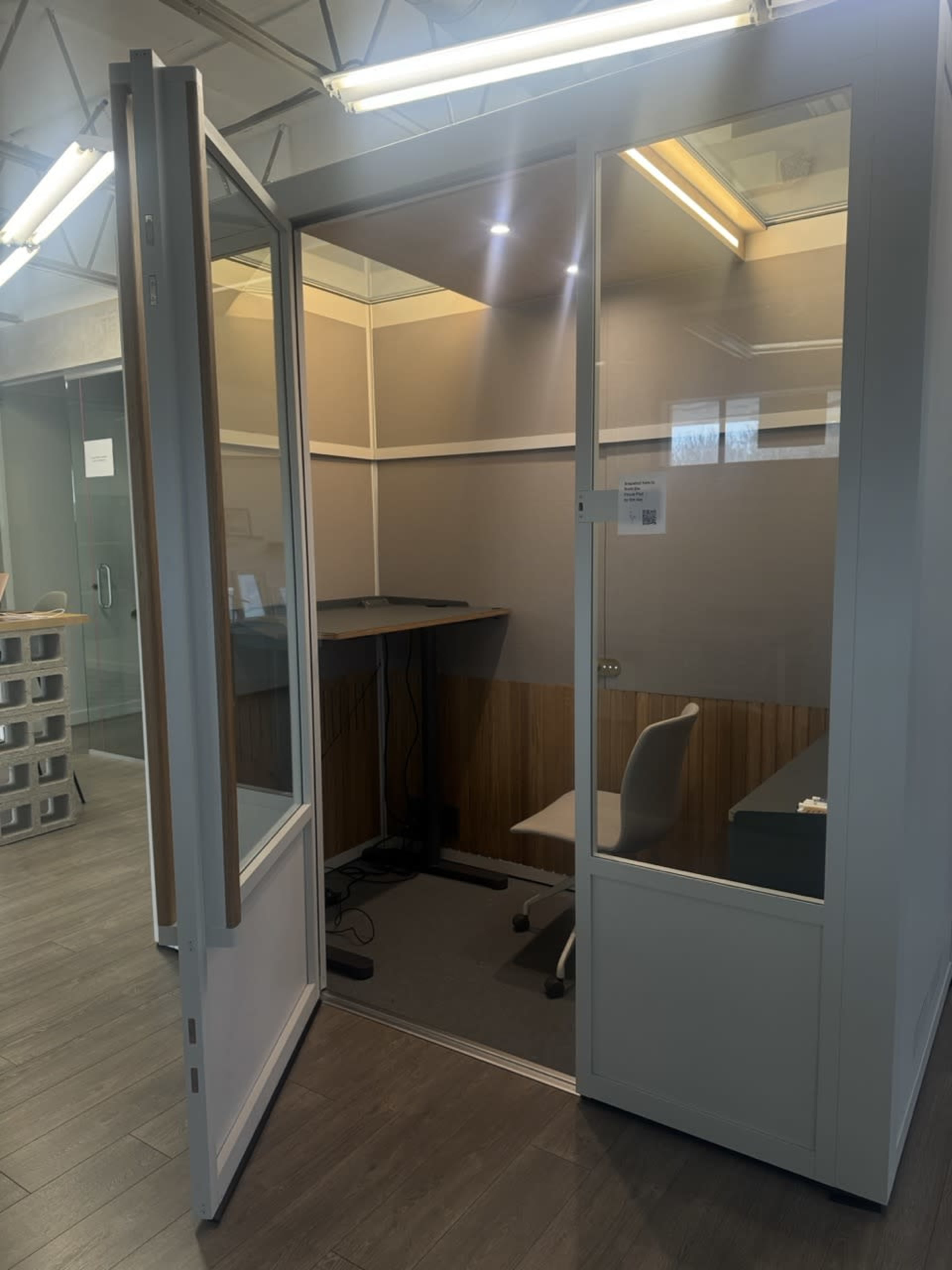The image shows a modern office phone booth with a glass door, a small desk, and a chair inside.