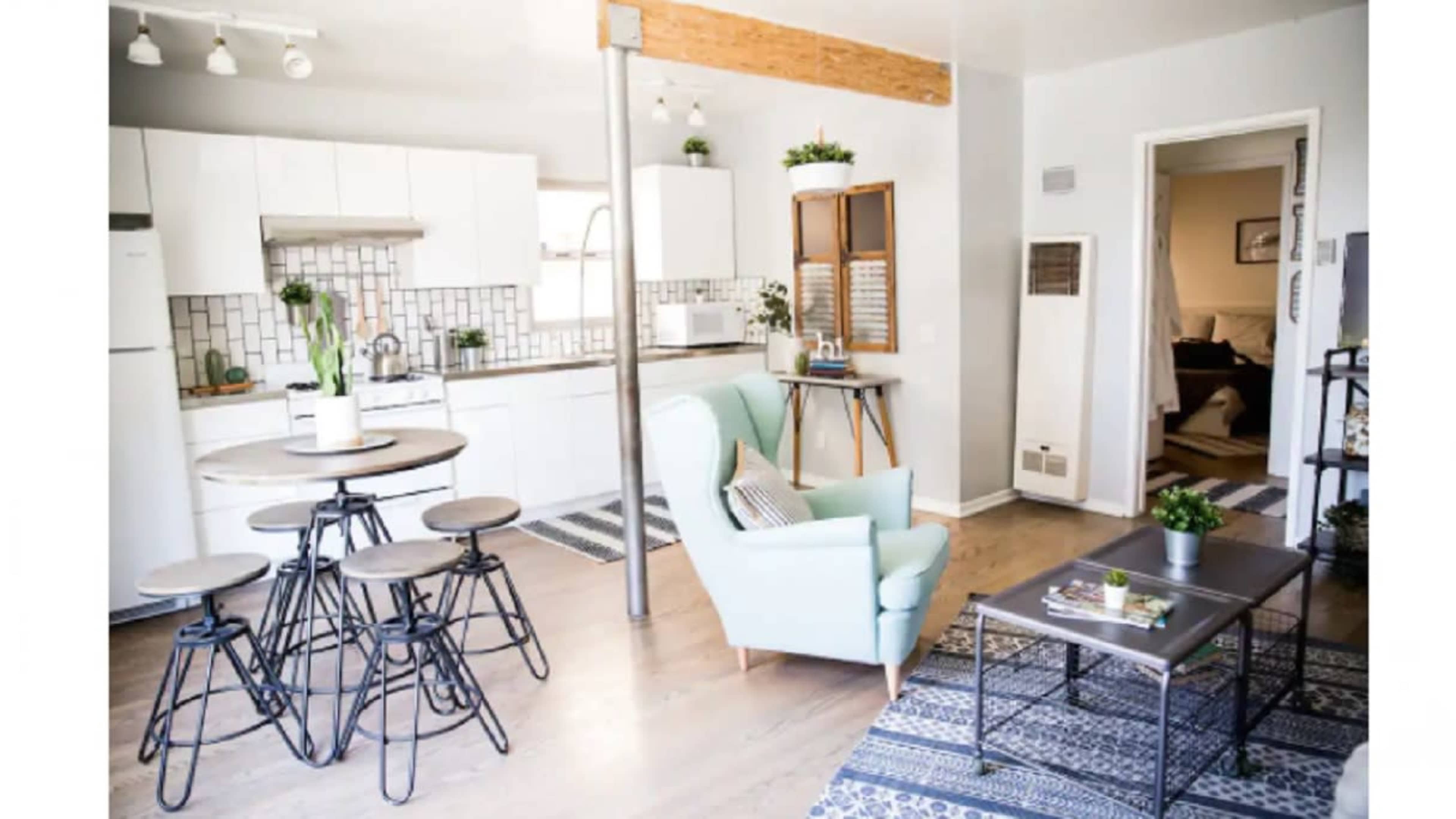 The image shows a modern kitchen and living area featuring a circular dining table with four stools, a green armchair, and a coffee table, all set against a backdrop of white cabinets and a patterned rug.