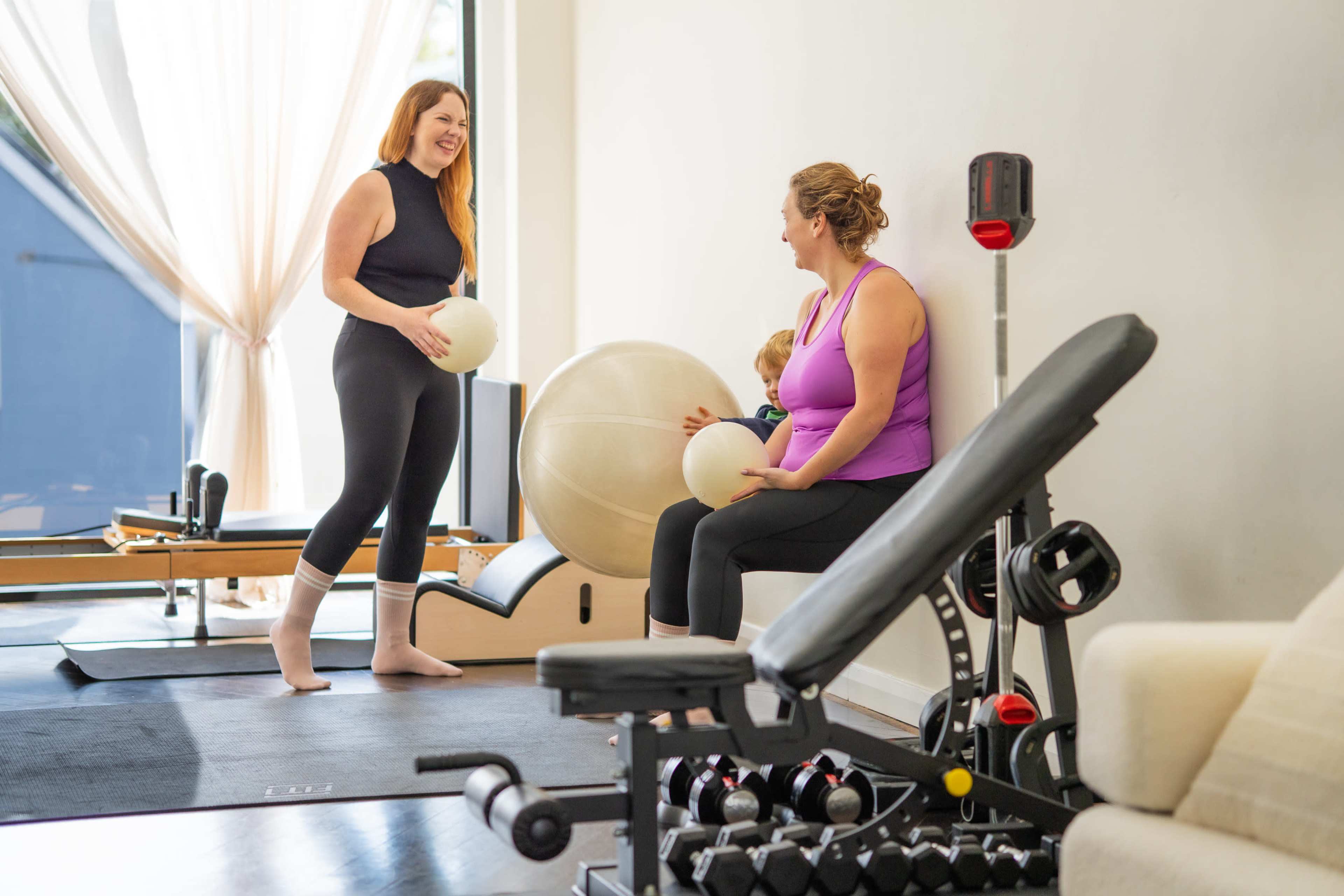 Two women are engaged in conversation while sitting and standing in a fitness studio, surrounded by exercise equipment and fitness balls.