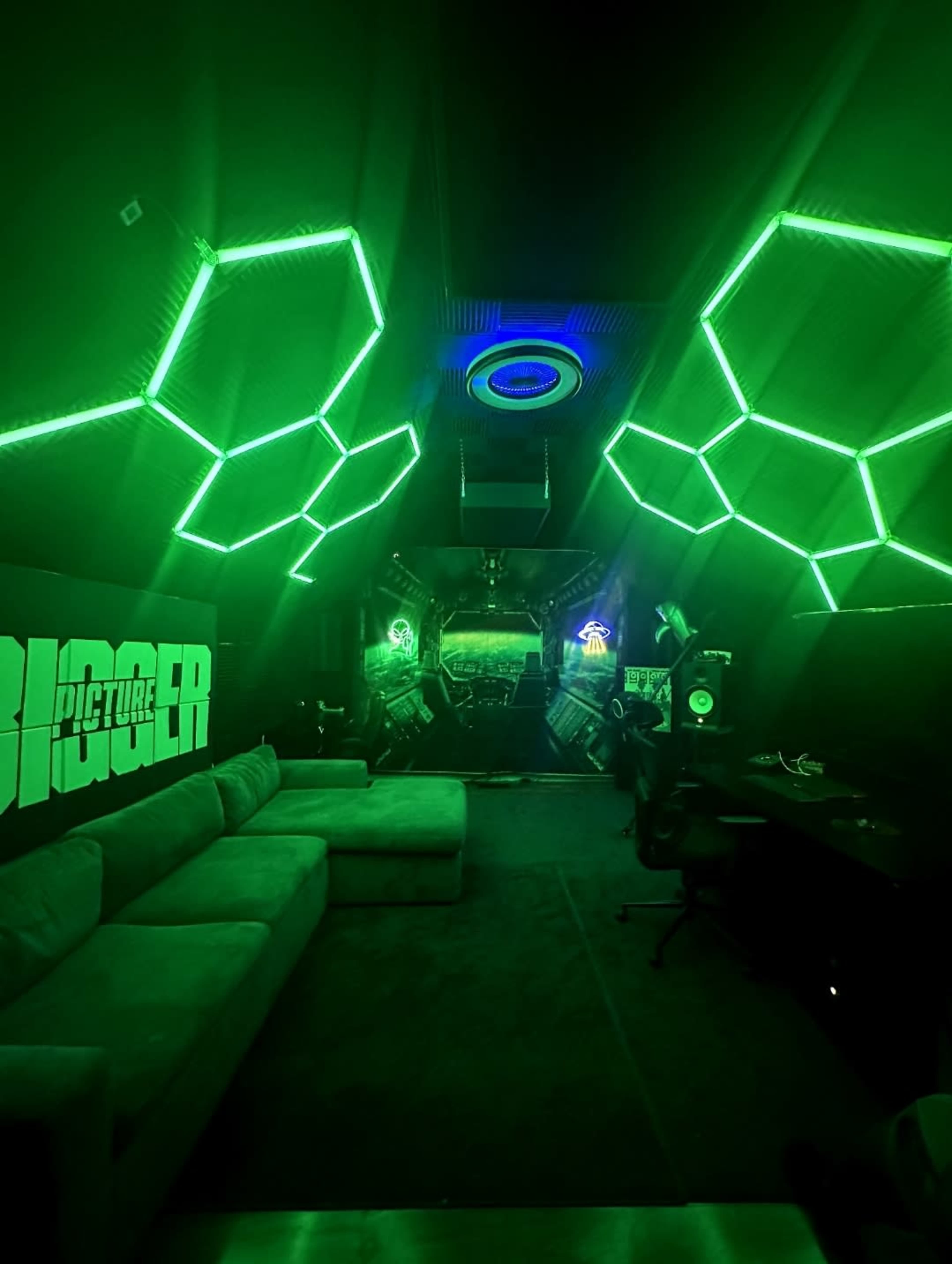 The image shows a dimly lit room with green LED lighting, featuring a couch, a workspace with audio equipment, and geometric wall designs.
