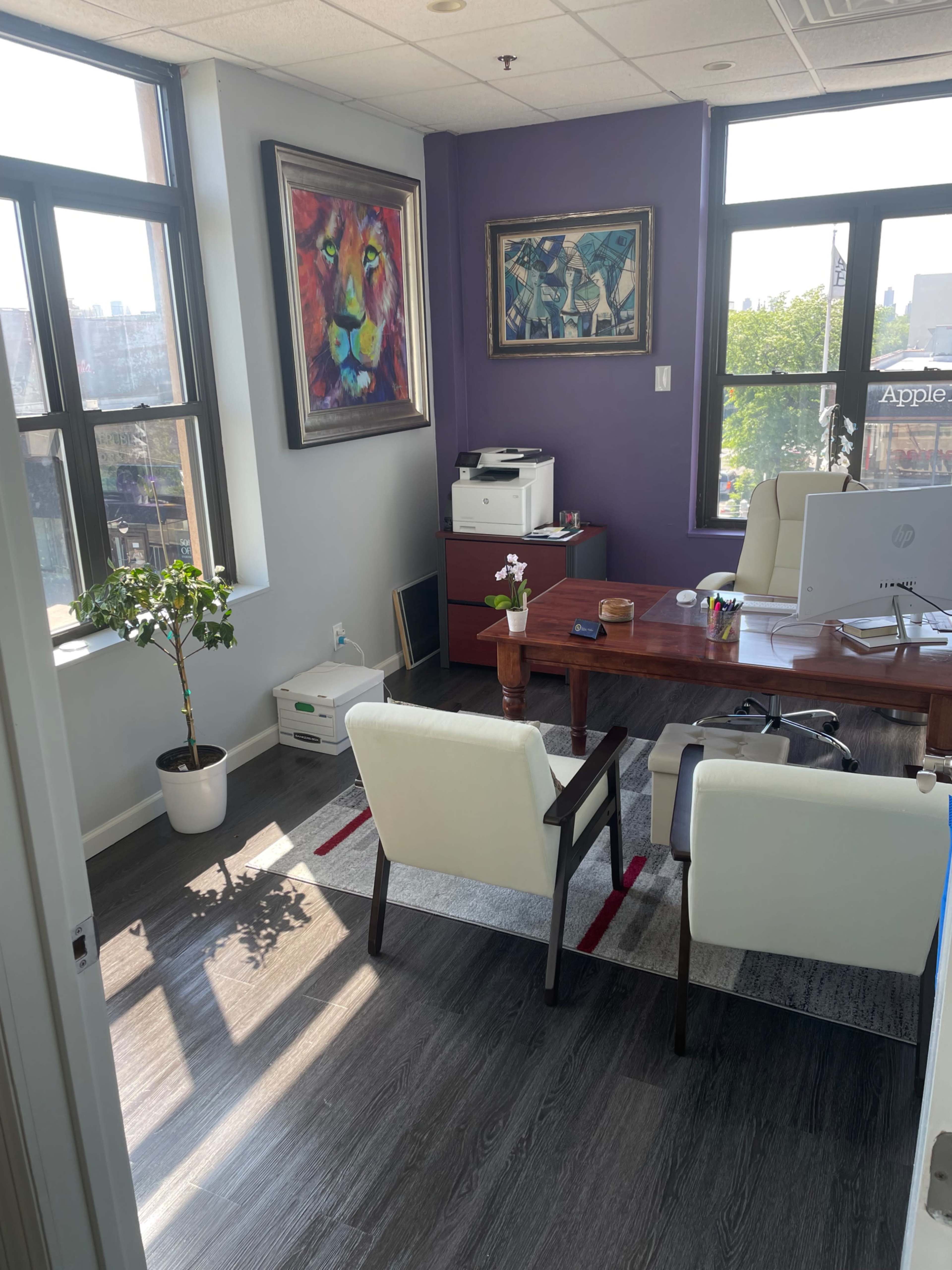 An office with a wooden desk, two white chairs, a potted plant, and artwork on the walls, featuring large windows that let in natural light.