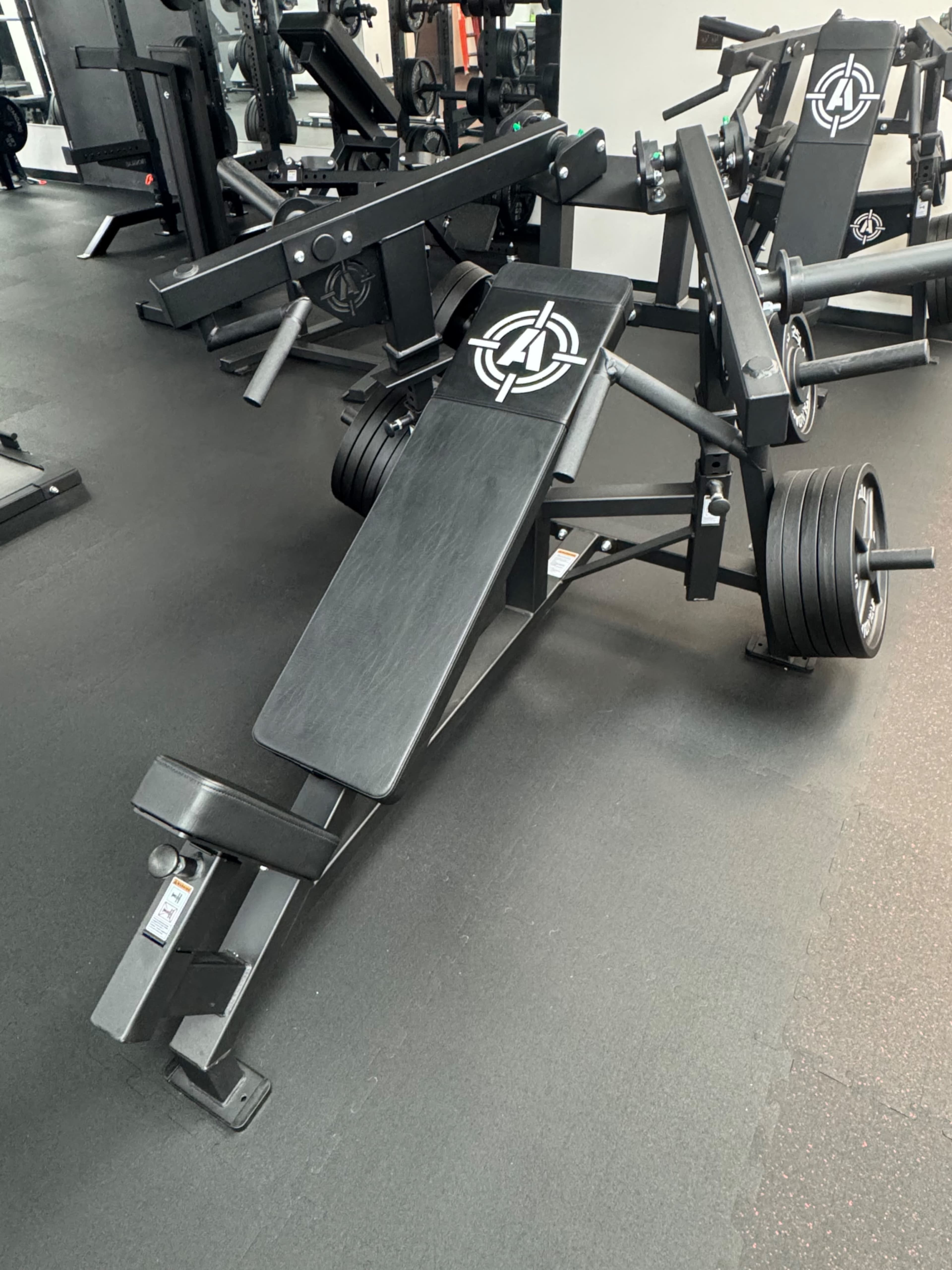 The image shows a weight-lifting bench with adjustable features and multiple weights on either side, situated in a gym environment.