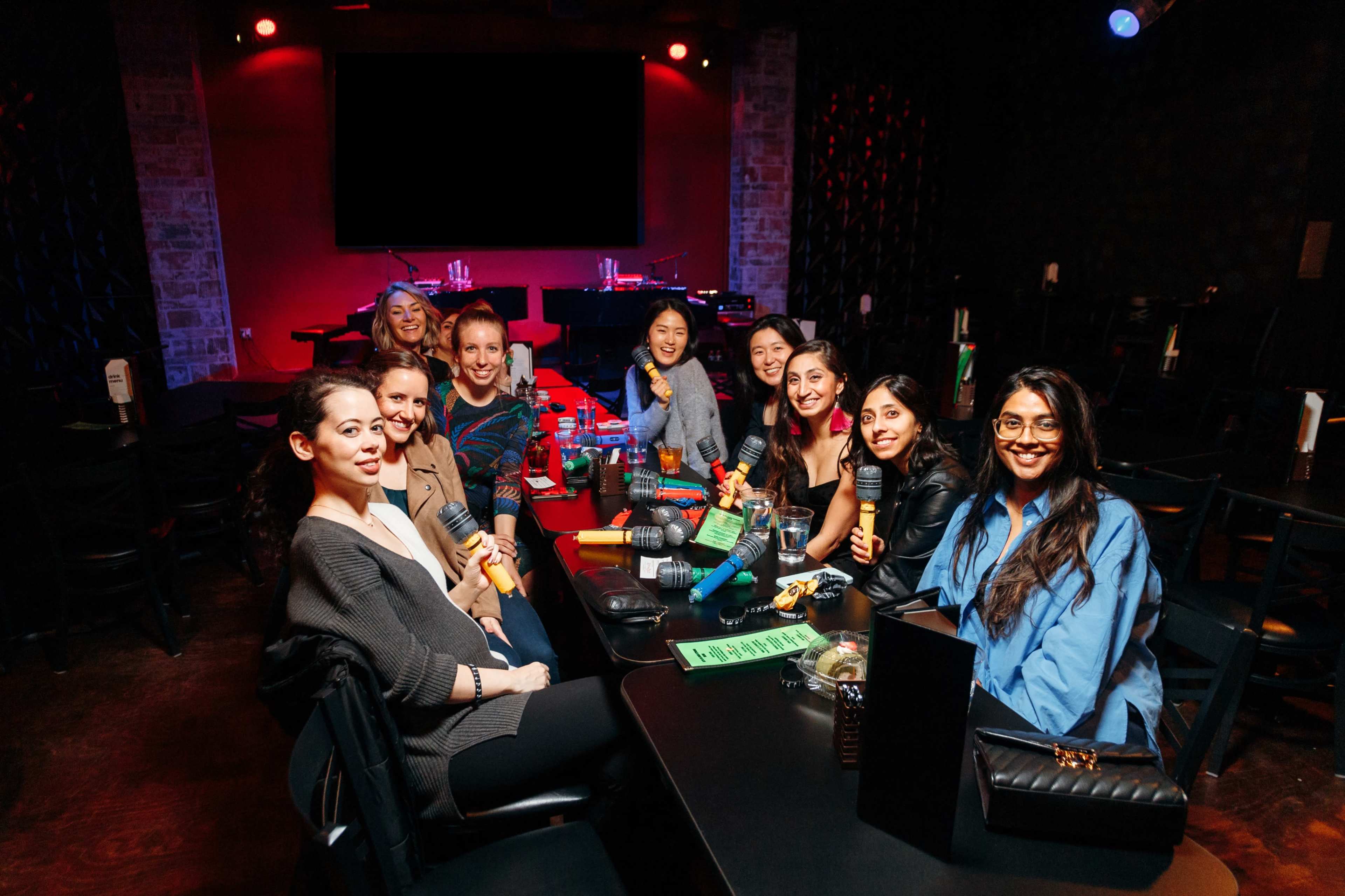 A group of nine women sits around a table in a dimly lit entertainment venue, each holding colorful microphones.