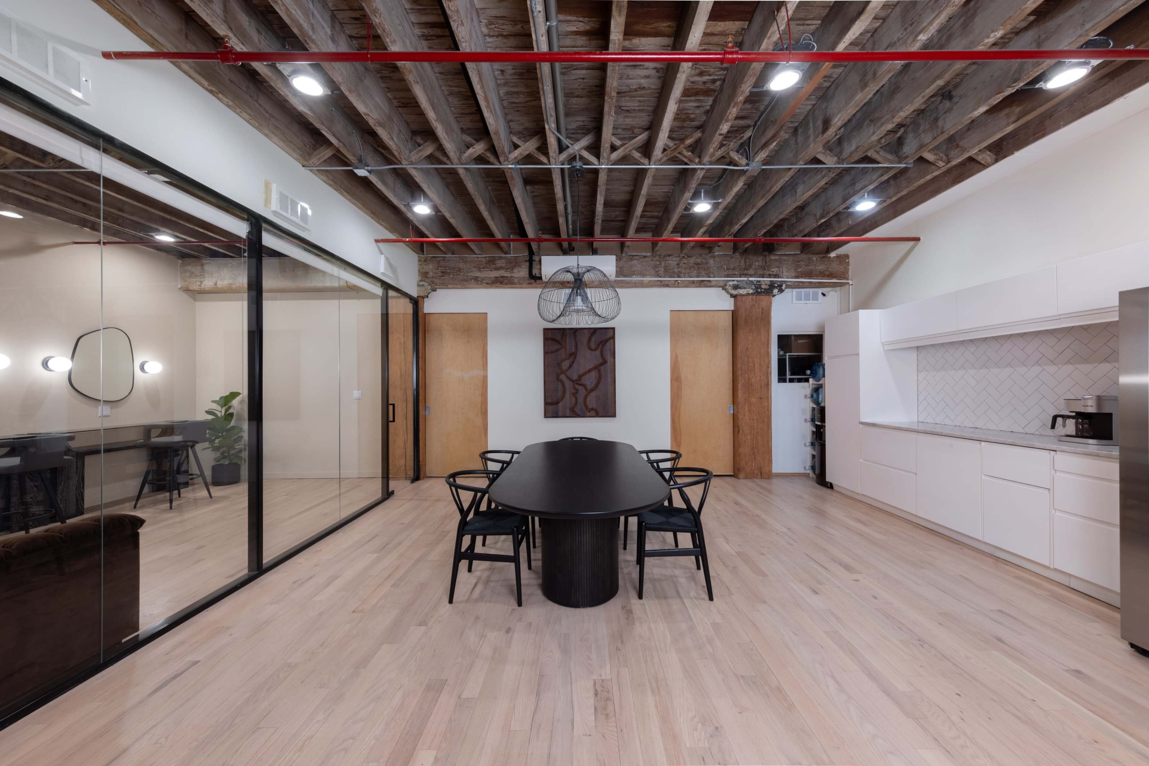 A modern conference room features a long black table surrounded by chairs, with exposed wooden beams and a glass wall leading to an adjacent space.