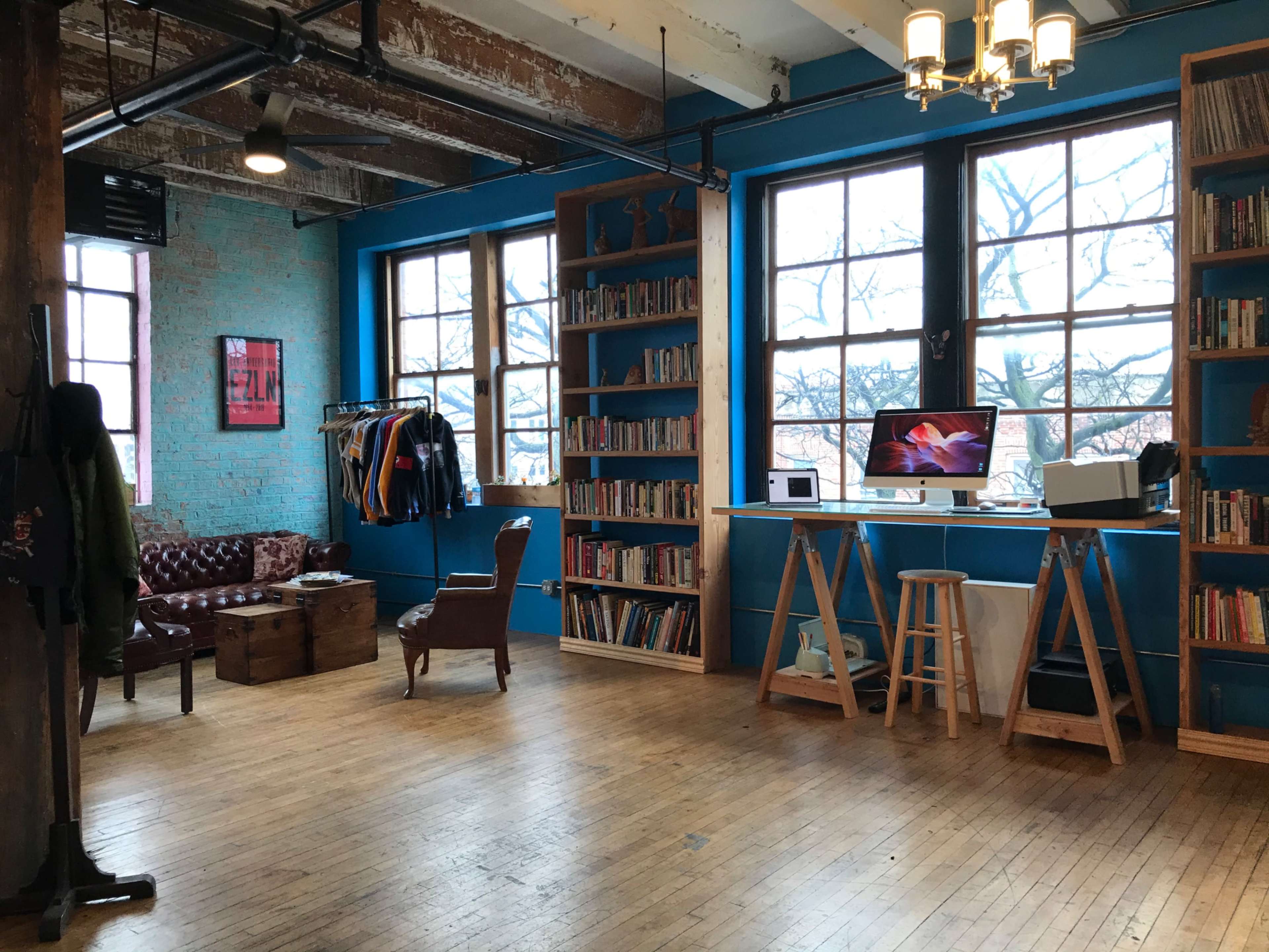 A spacious room with blue walls, wooden flooring, large windows, a bookshelf filled with books, a vintage armchair, and a computer setup on a desk.