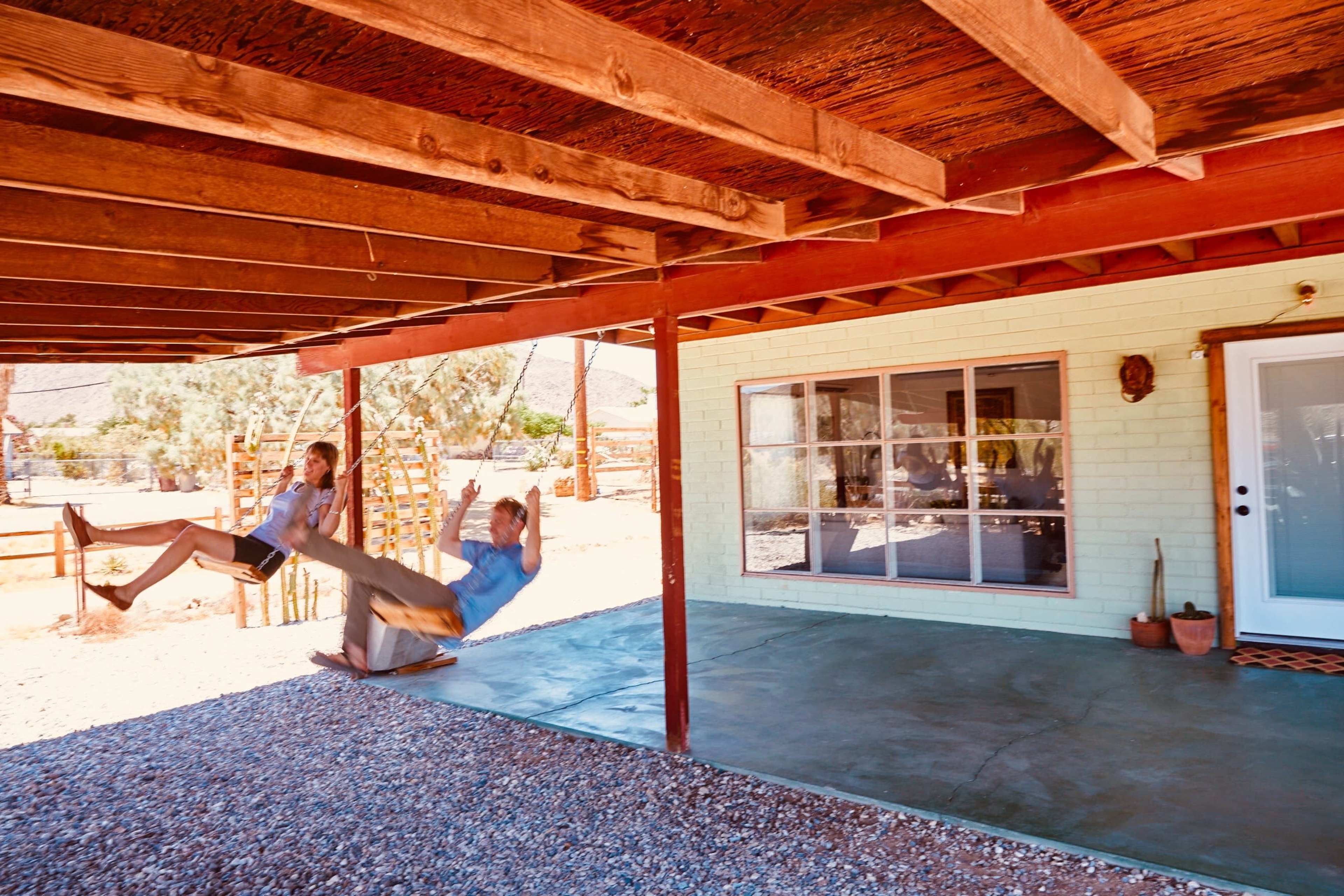 Two children are swinging on a rope swing beneath a wooden porch, with a desert landscape visible in the background.