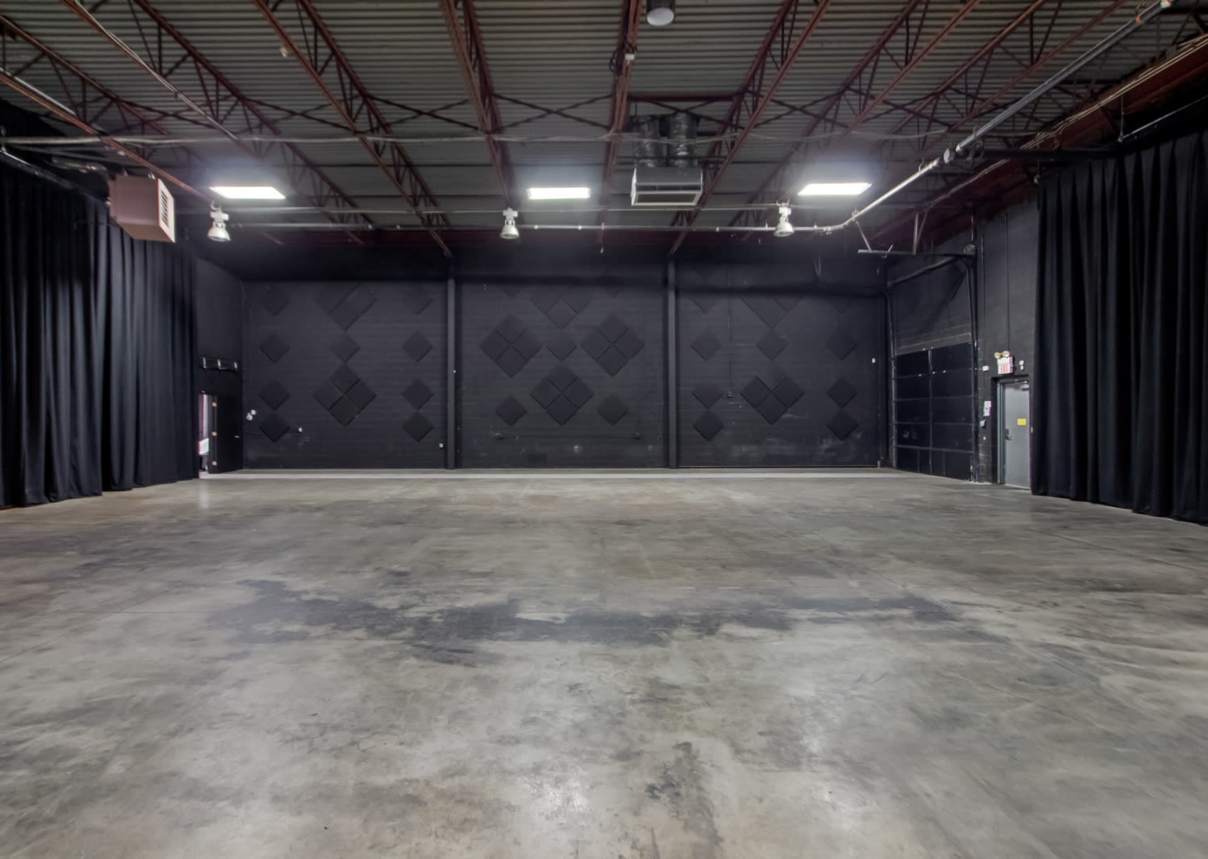 A large empty room with a concrete floor, black walls, and black curtains, illuminated by overhead lights.