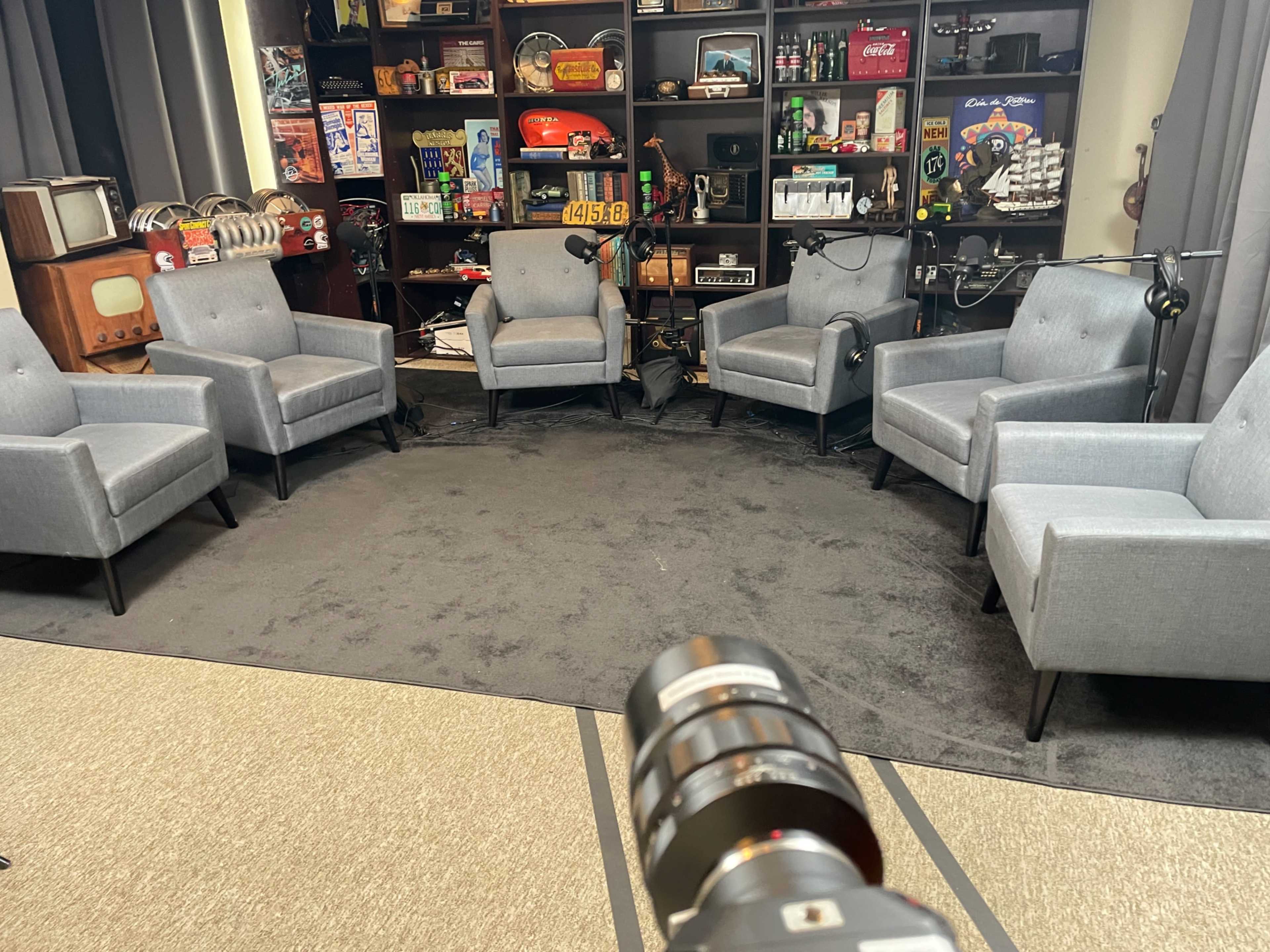 The image shows a neatly arranged circle of four gray armchairs on a dark rug, facing each other in a studio setting, with various items displayed on shelves in the background.