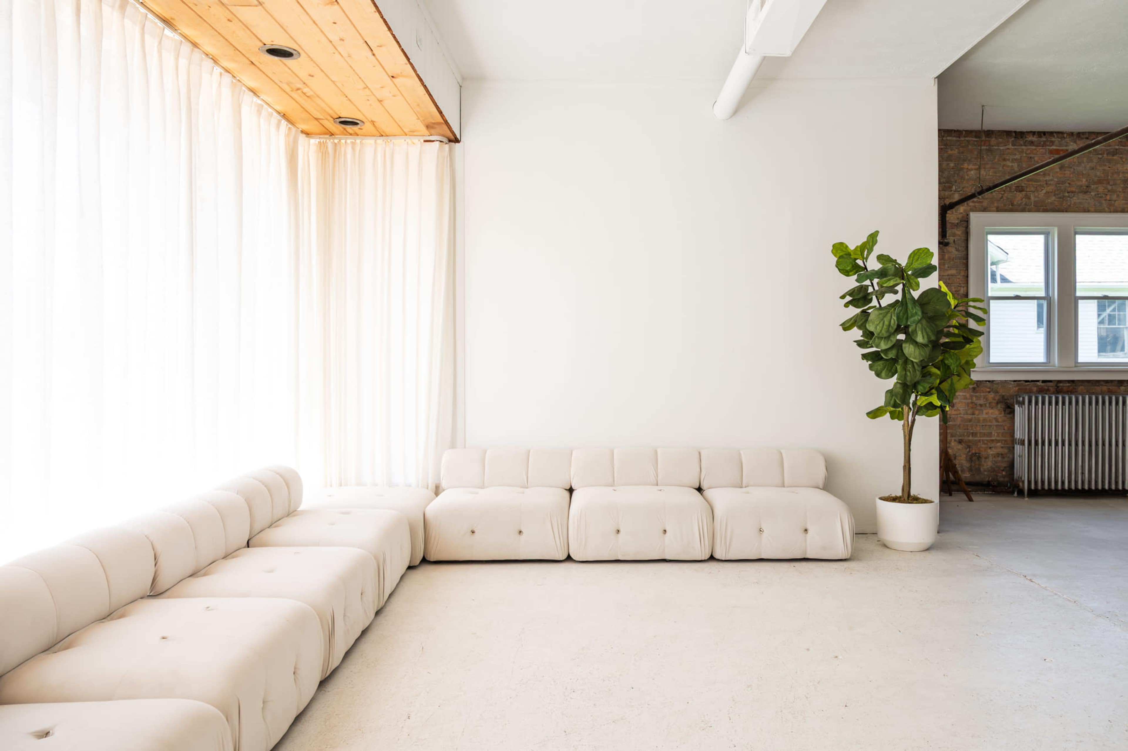 The image shows a minimalist interior space featuring a light-colored upholstered sofa, large windows with sheer curtains, and a potted plant in the corner.