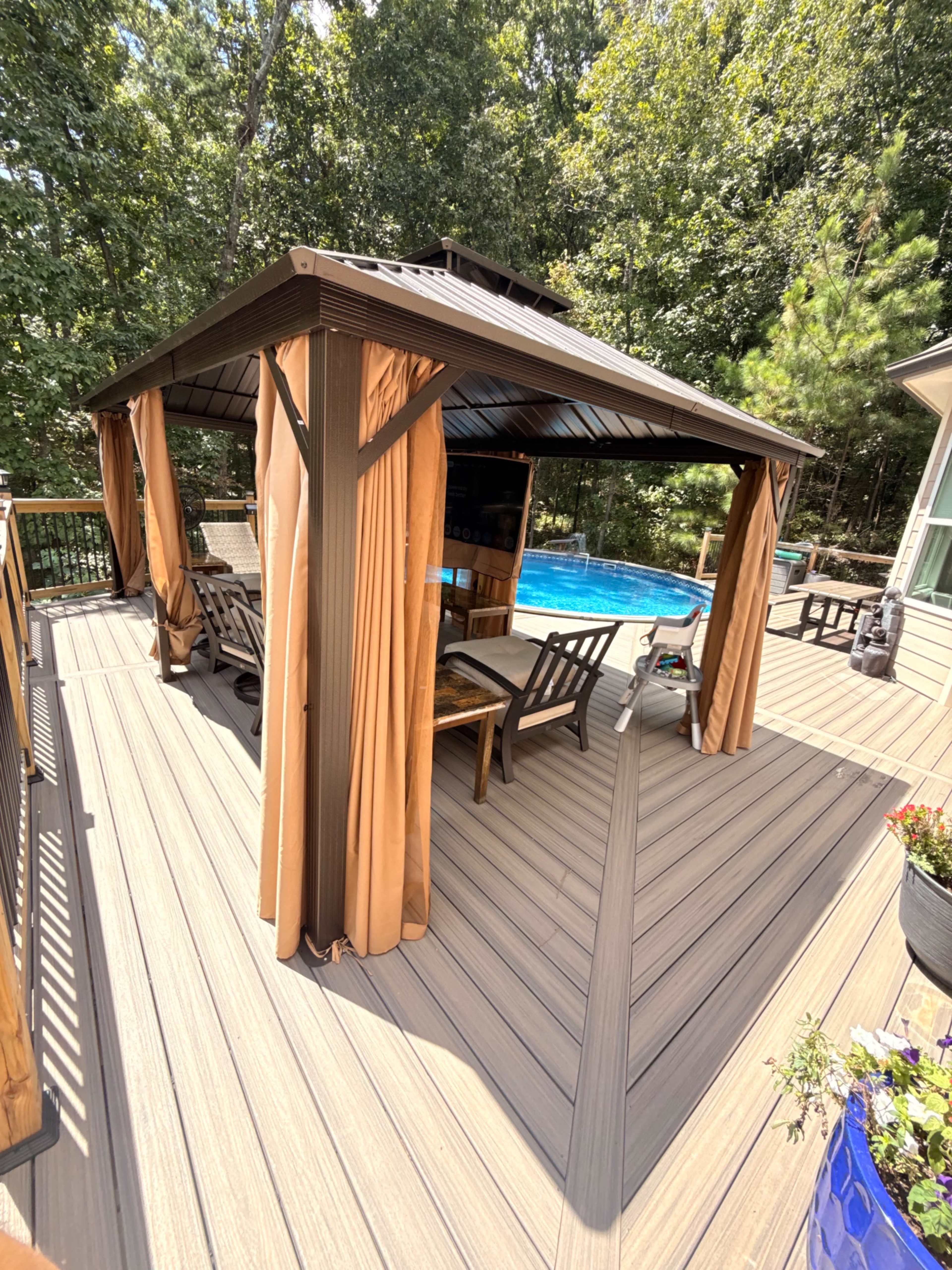 The image shows a shaded gazebo on a wooden deck adjacent to a swimming pool, surrounded by trees.