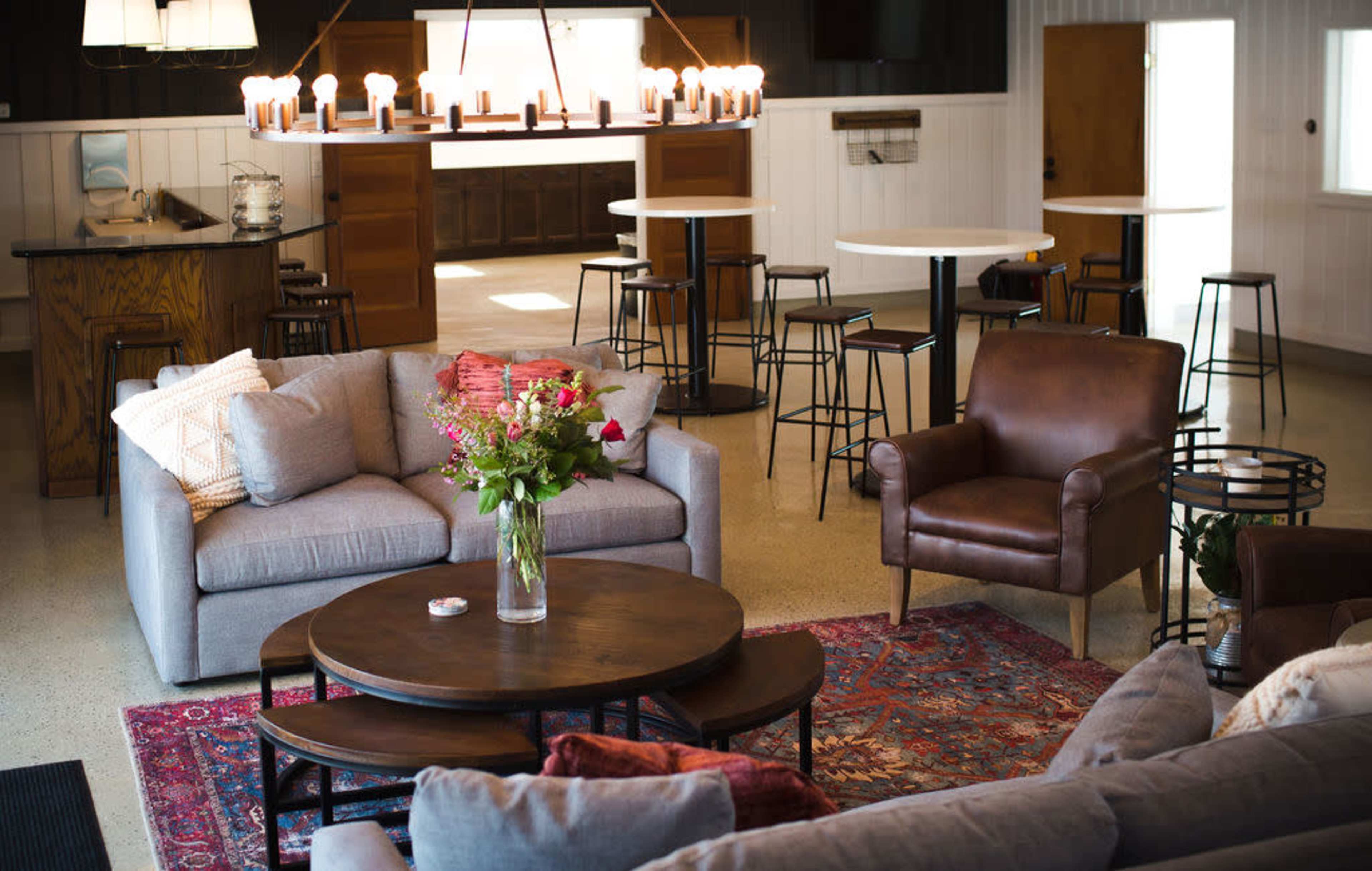 The image shows a modern interior space with a seating area featuring two sofas, a round coffee table, and a floral centerpiece, along with a dining area containing high tables and chairs.