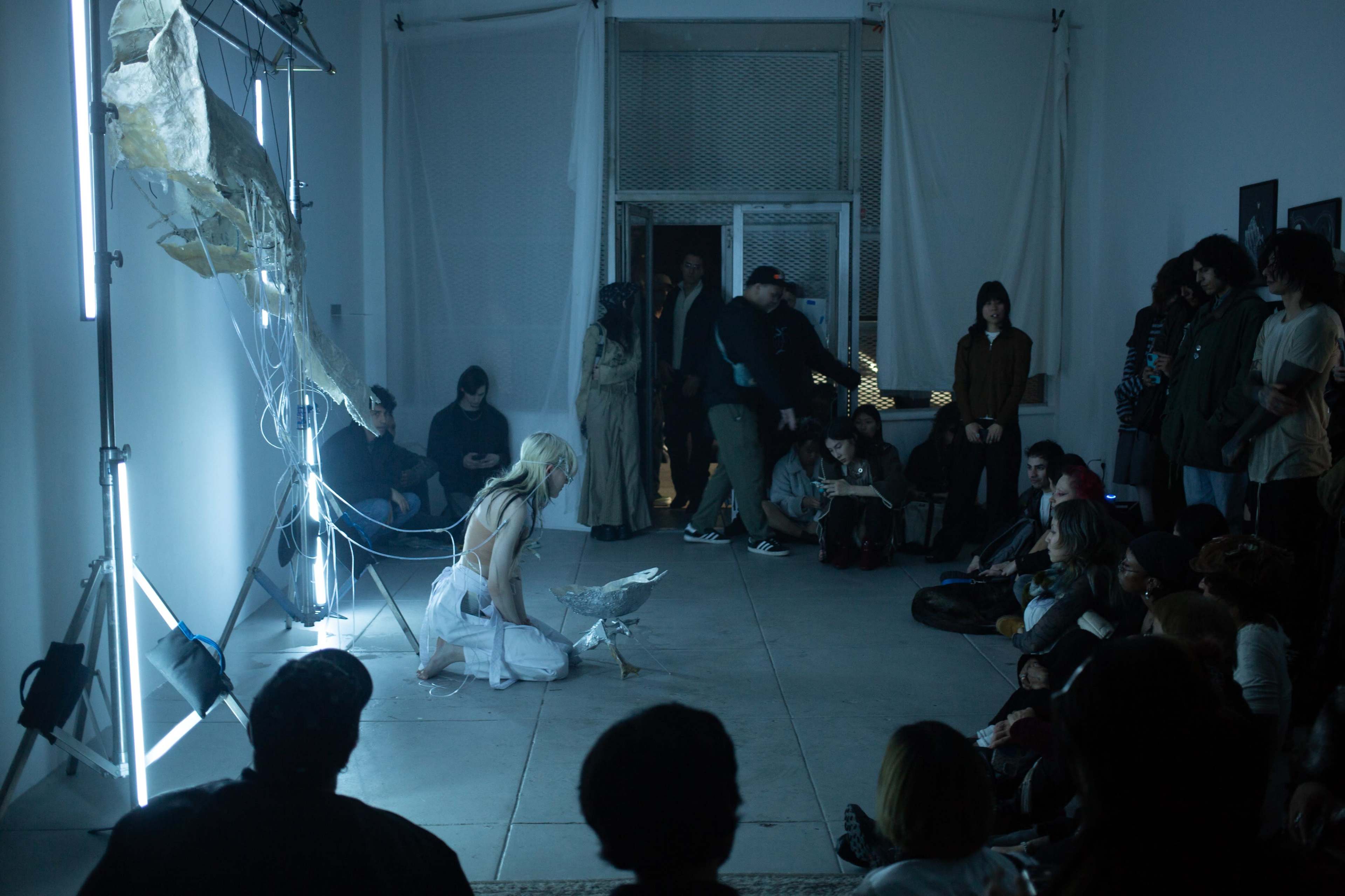 A performer dressed in white interacts with a sculptural installation in a gallery space, while an audience sits on the floor, watching attentively.