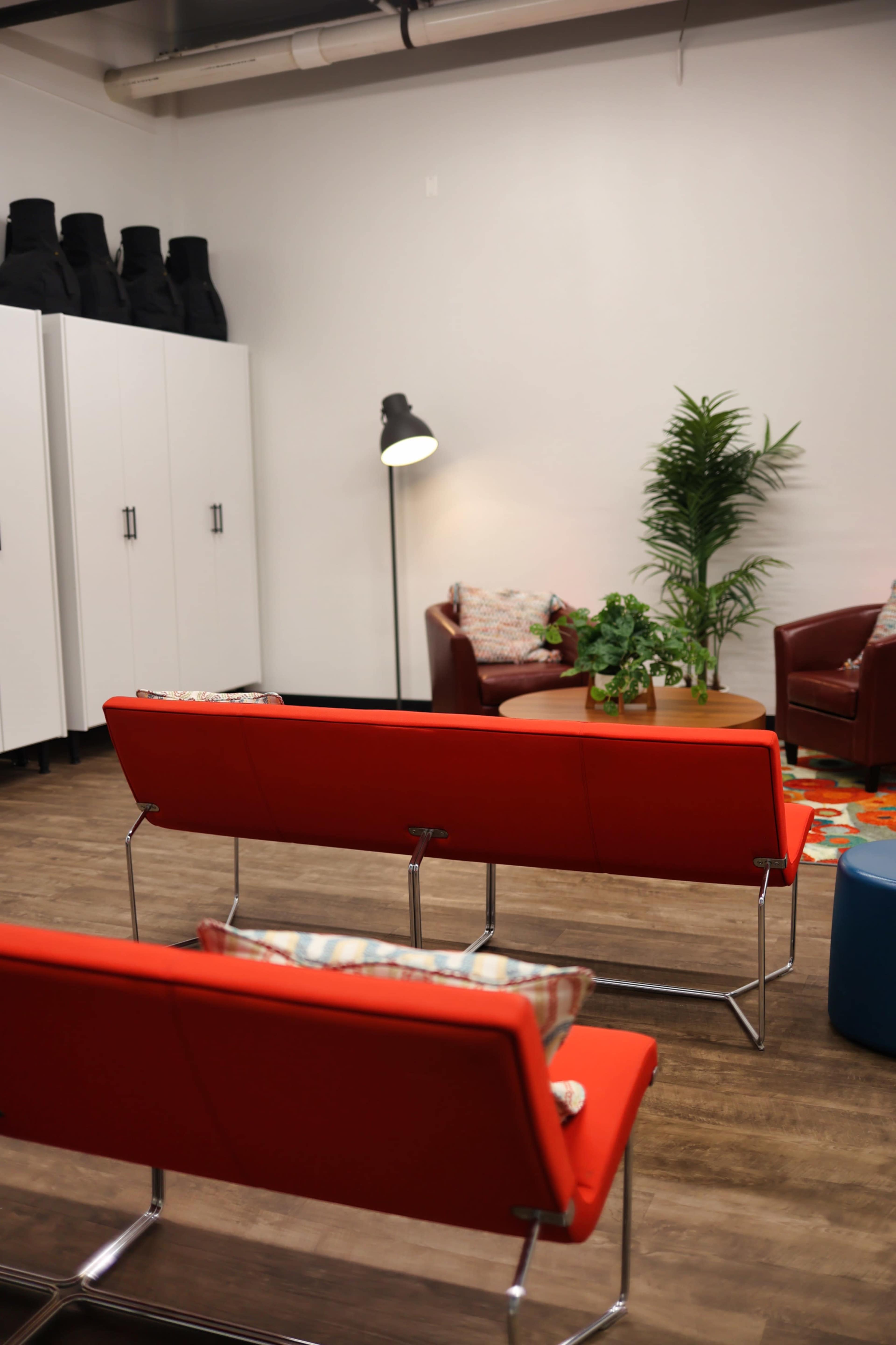 The scene features a modern lounge area with red seating, a round table, and a potted plant, set against a backdrop of white cabinets and a floor lamp.