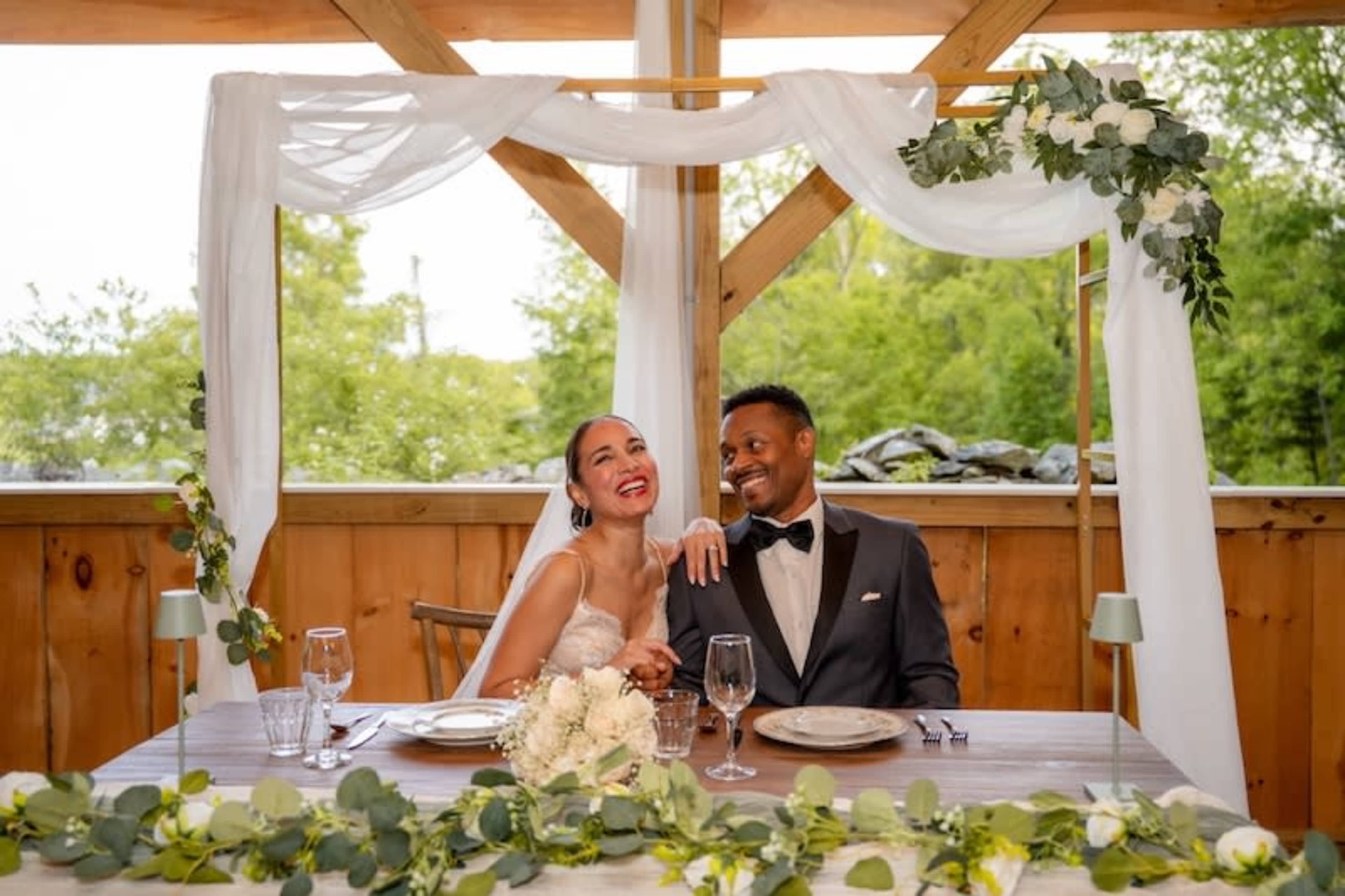 A newlywed couple smiles together at a decorated wedding table under a floral arch.