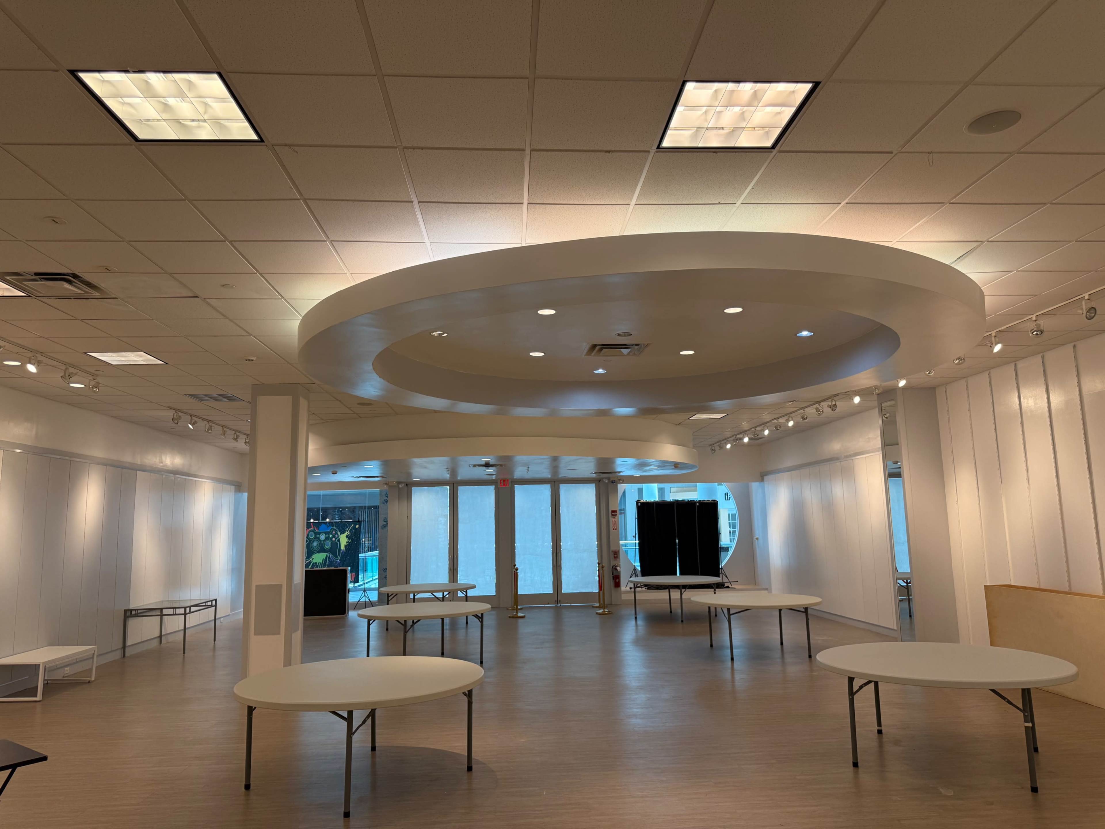 The image shows a spacious room with white walls, multiple round tables set up, and a ceiling featuring circular designs and recessed lighting.