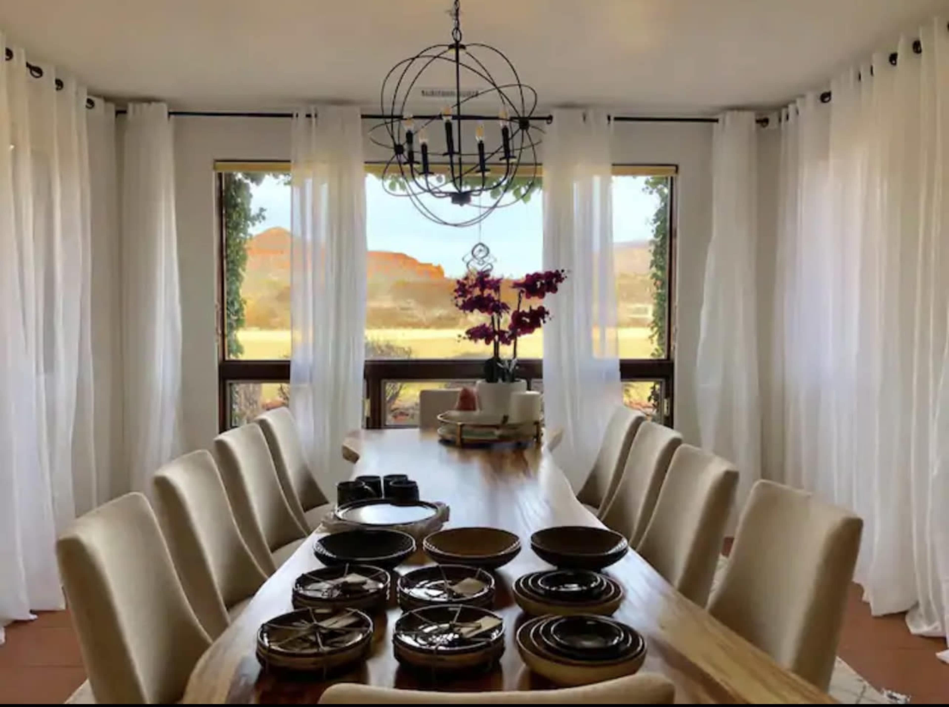 A large dining table is set with dinnerware, surrounded by upholstered chairs, and features large windows with sheer curtains overlooking a landscape with distant hills.