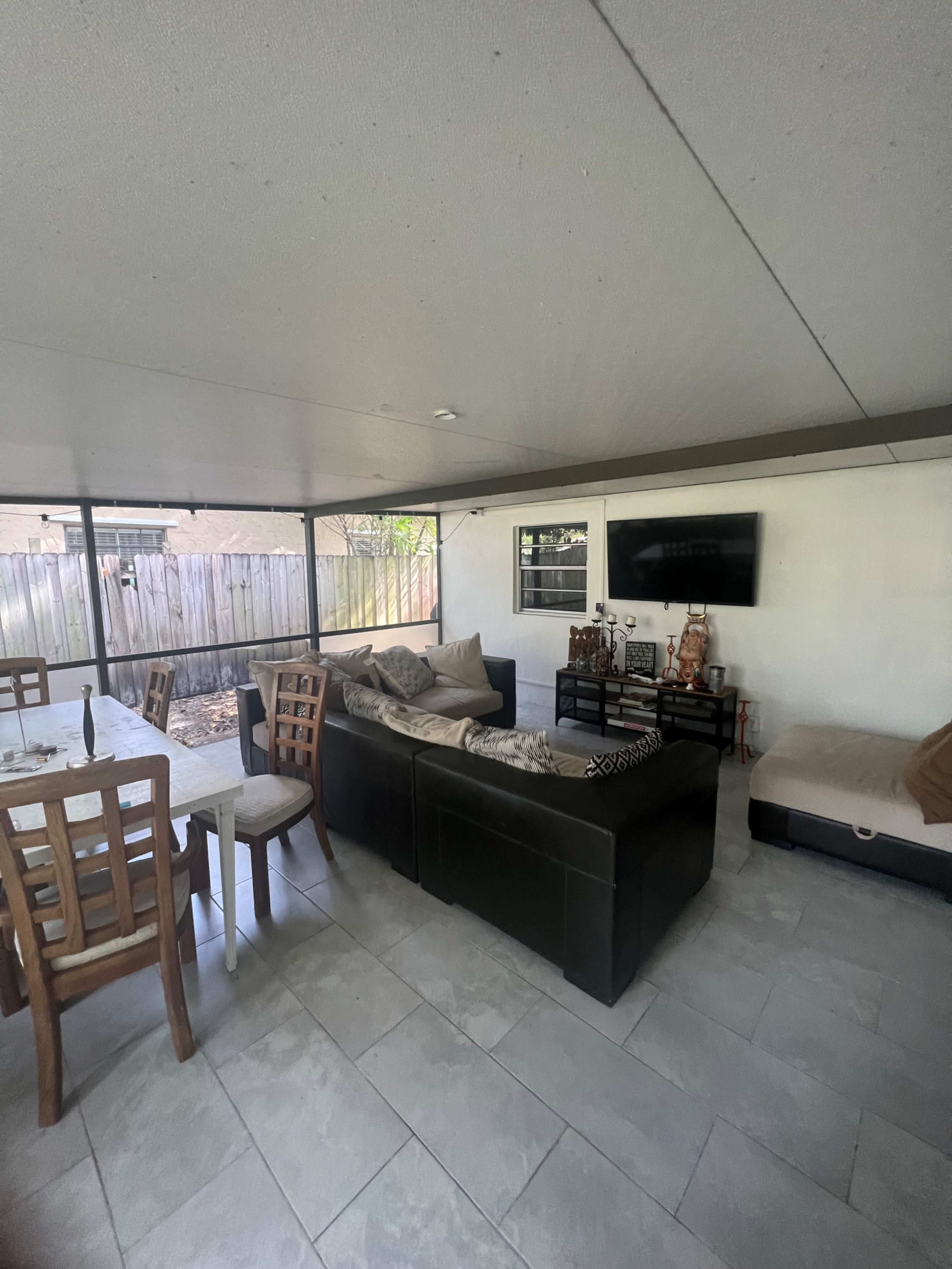A covered outdoor living space featuring a dining table and chairs, a sofa with cushions, and a television mounted on the wall.