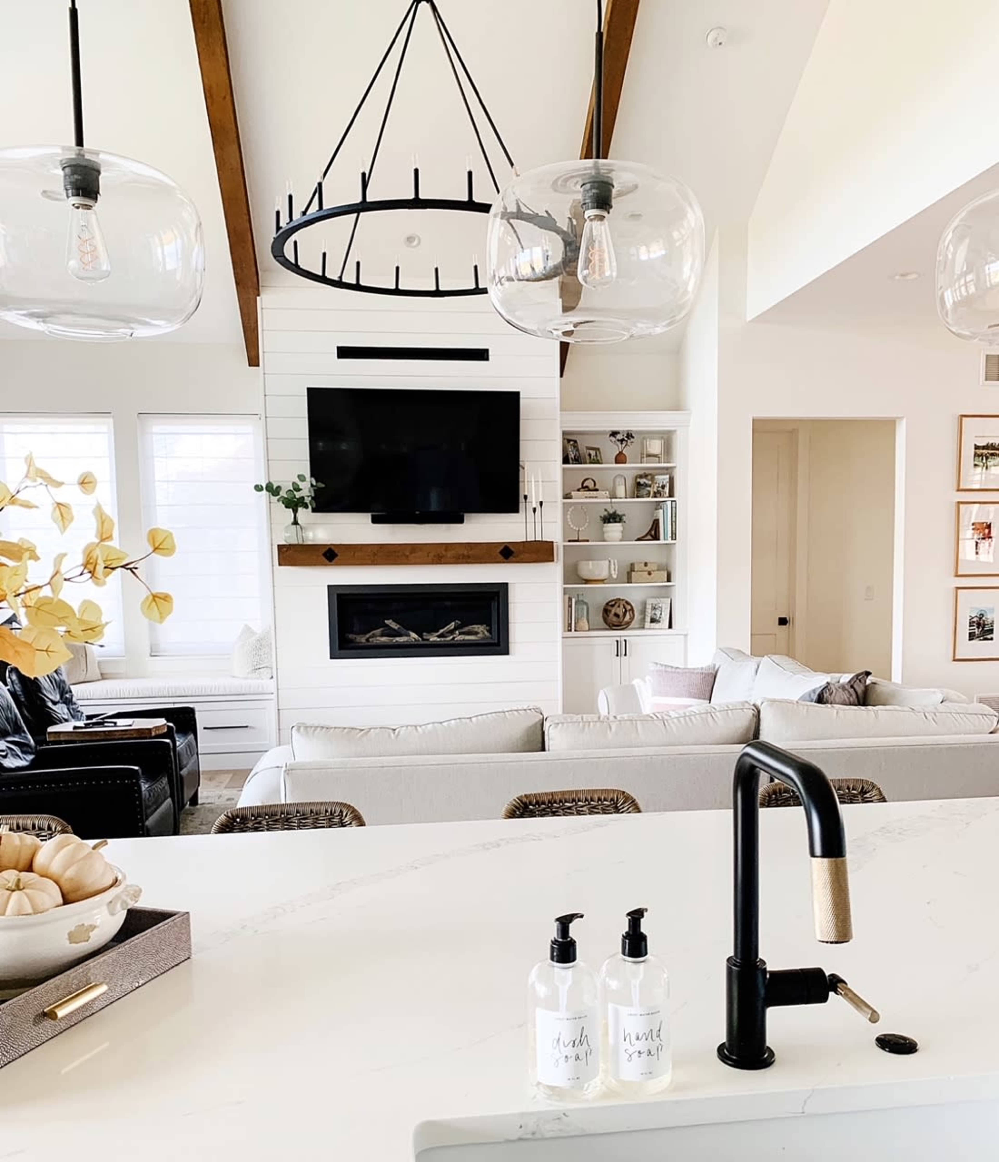 A modern living room featuring a white sectional sofa, a wall-mounted television above a fireplace, and a kitchen area with a black faucet and soap dispensers on the counter.