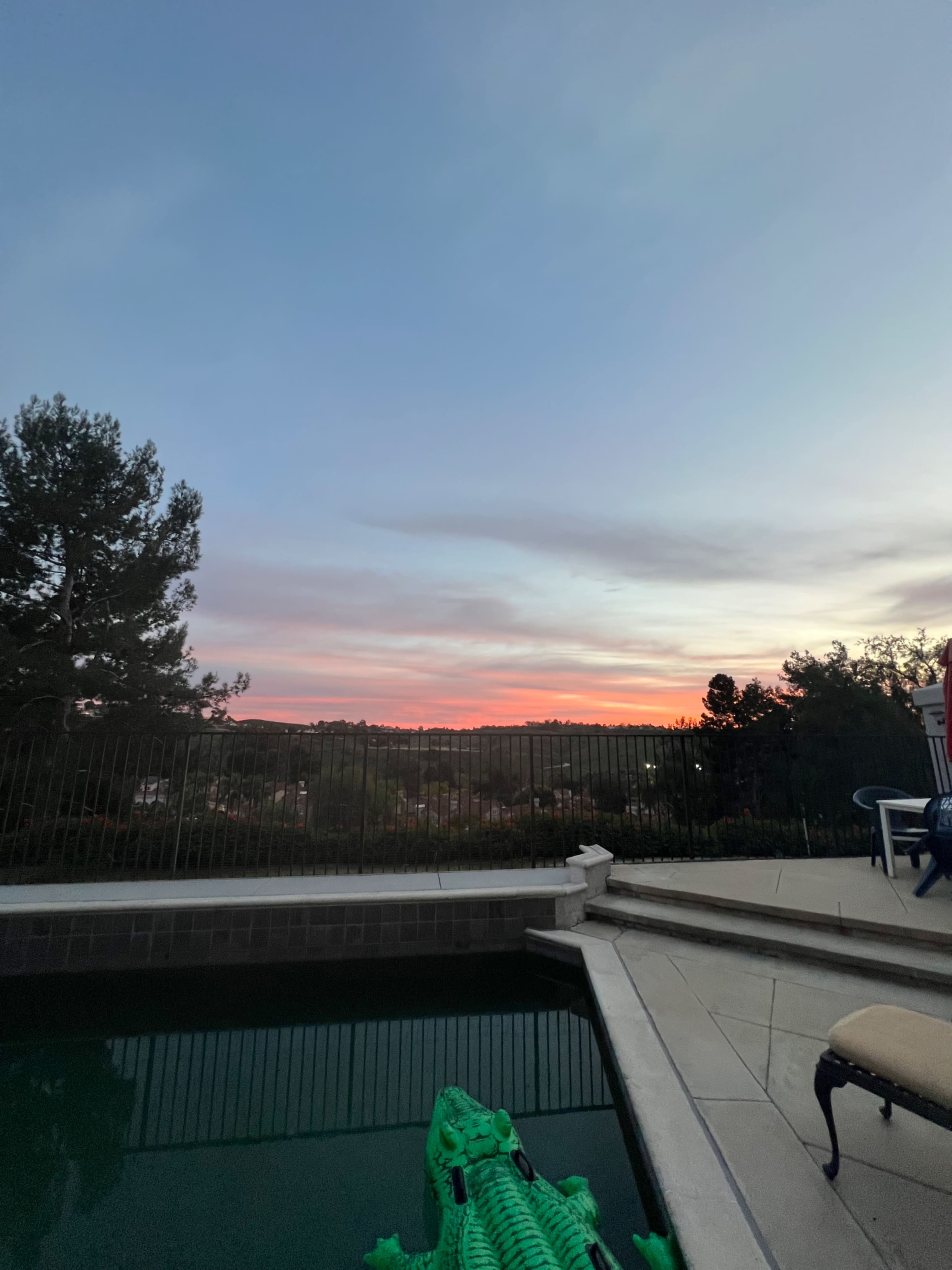 A poolside view at dusk with a green inflatable crocodile floating in the water and a sunset illuminating the sky.