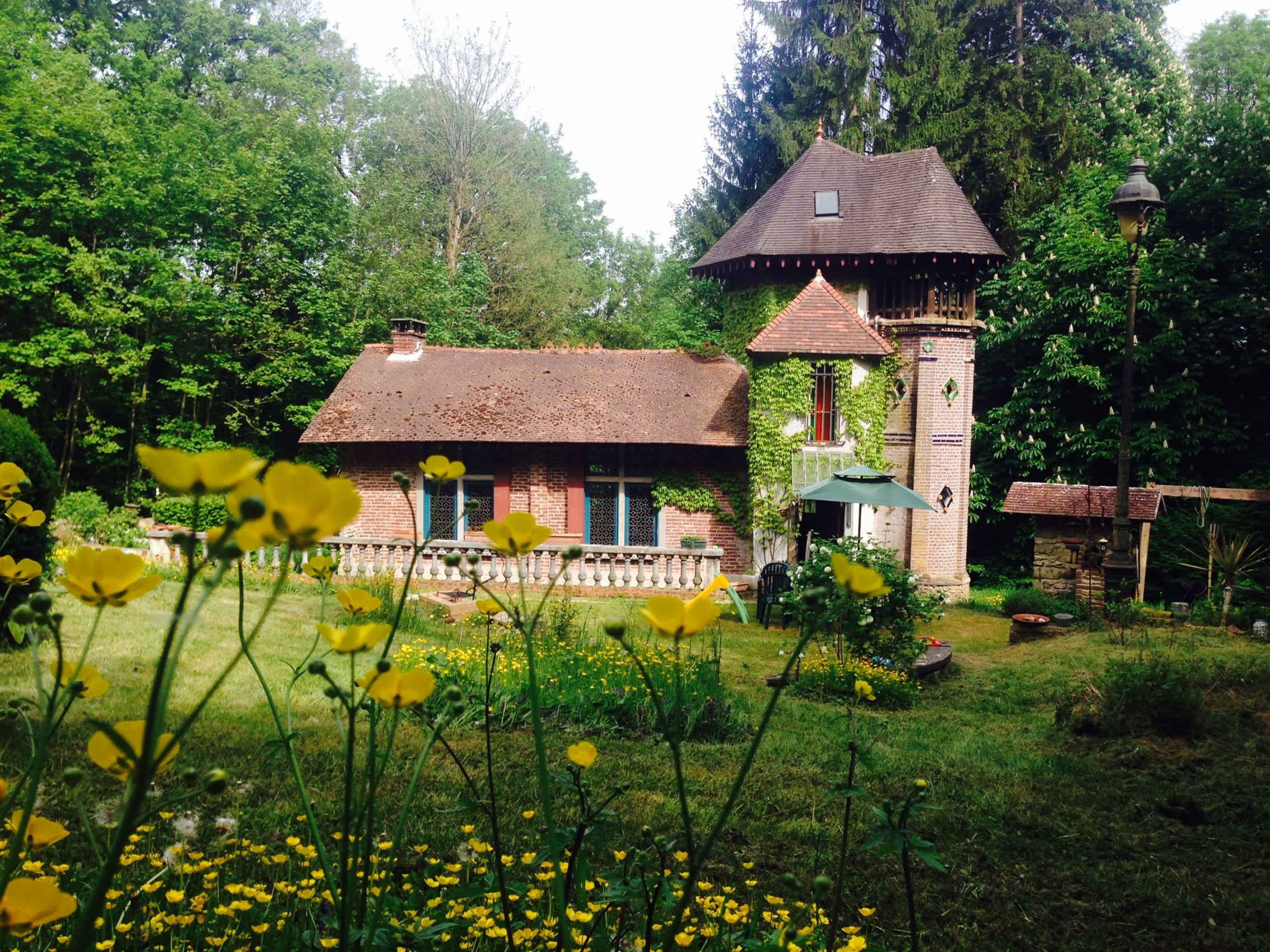A brick house with a tower is surrounded by green trees and colorful flowers in a garden.