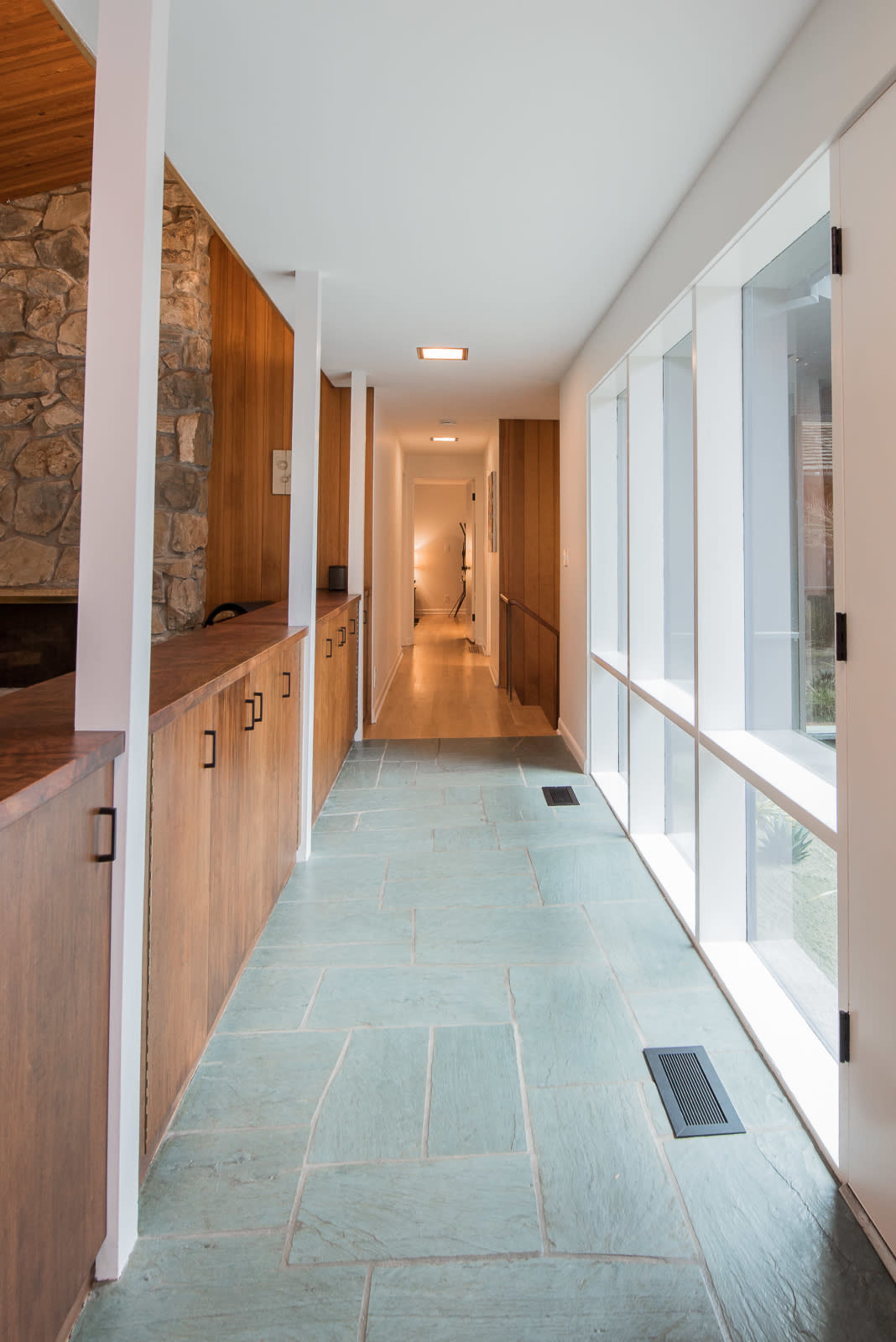 A long, modern hallway featuring stone flooring and wooden cabinets along one side, with large windows letting in light.