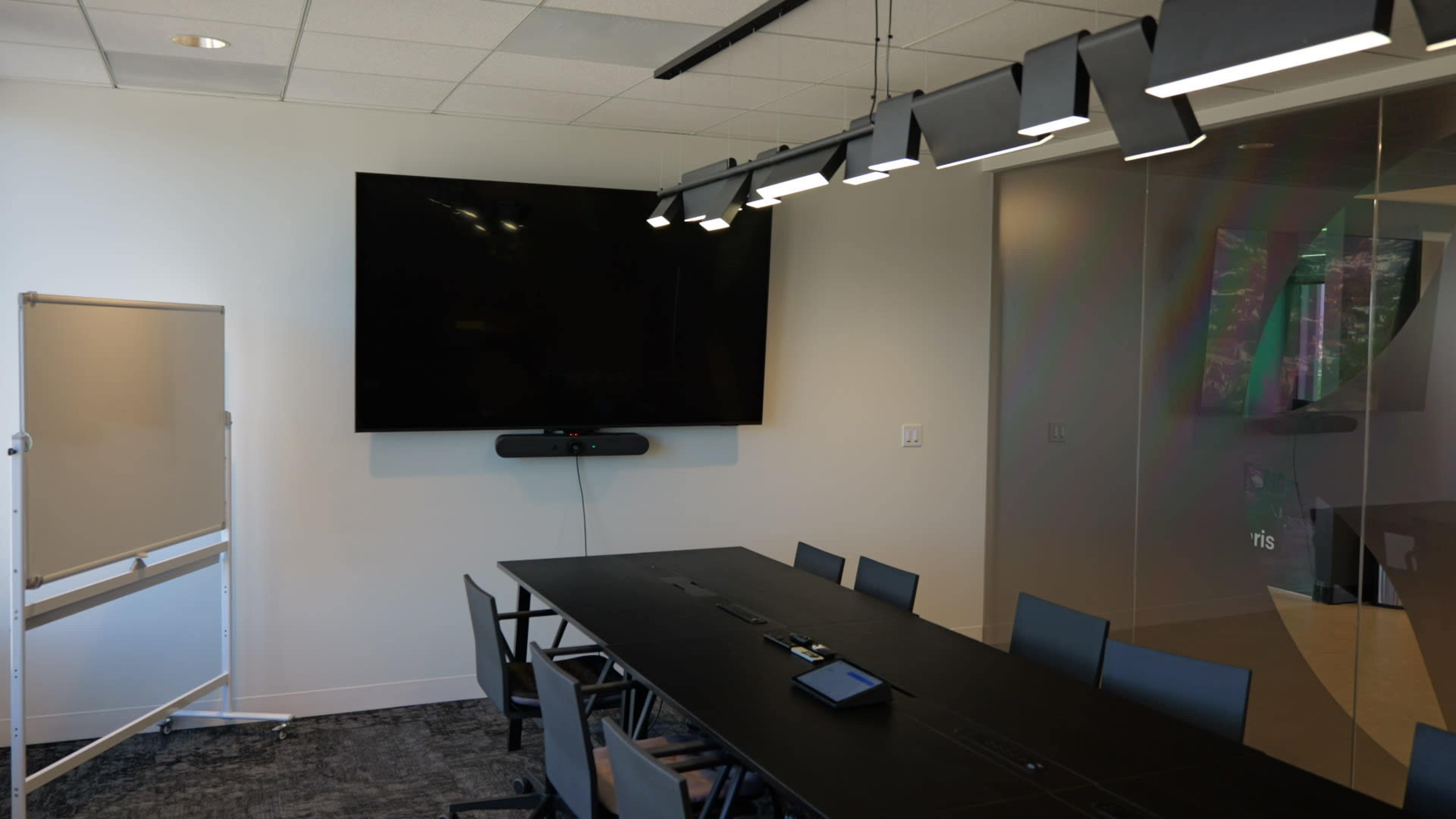 A modern conference room features a large flat-screen TV mounted on the wall, a long table with chairs, and a whiteboard in the corner.