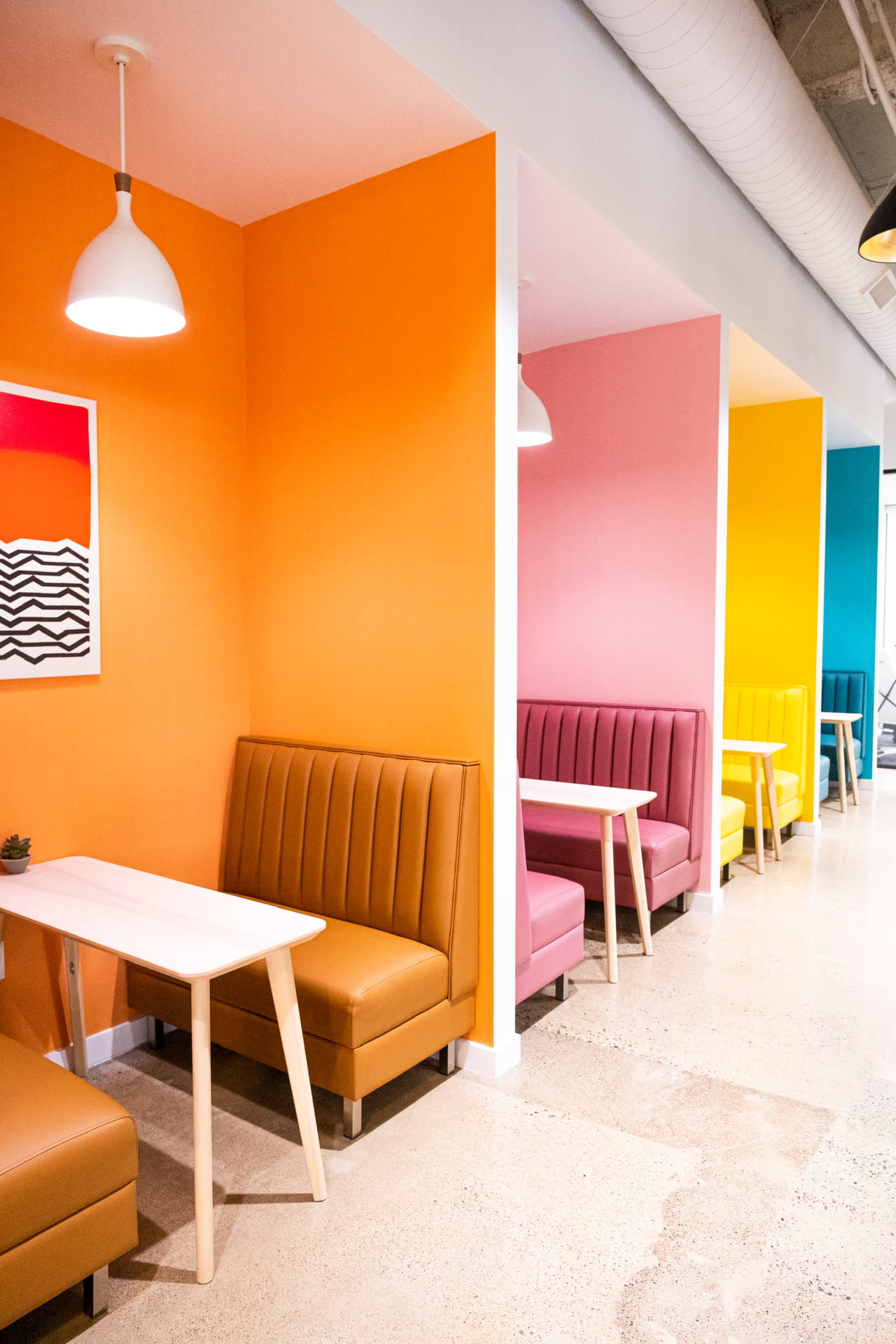 The image shows a modern café interior featuring colorful booth seating in orange, pink, and yellow, with light wood tables.