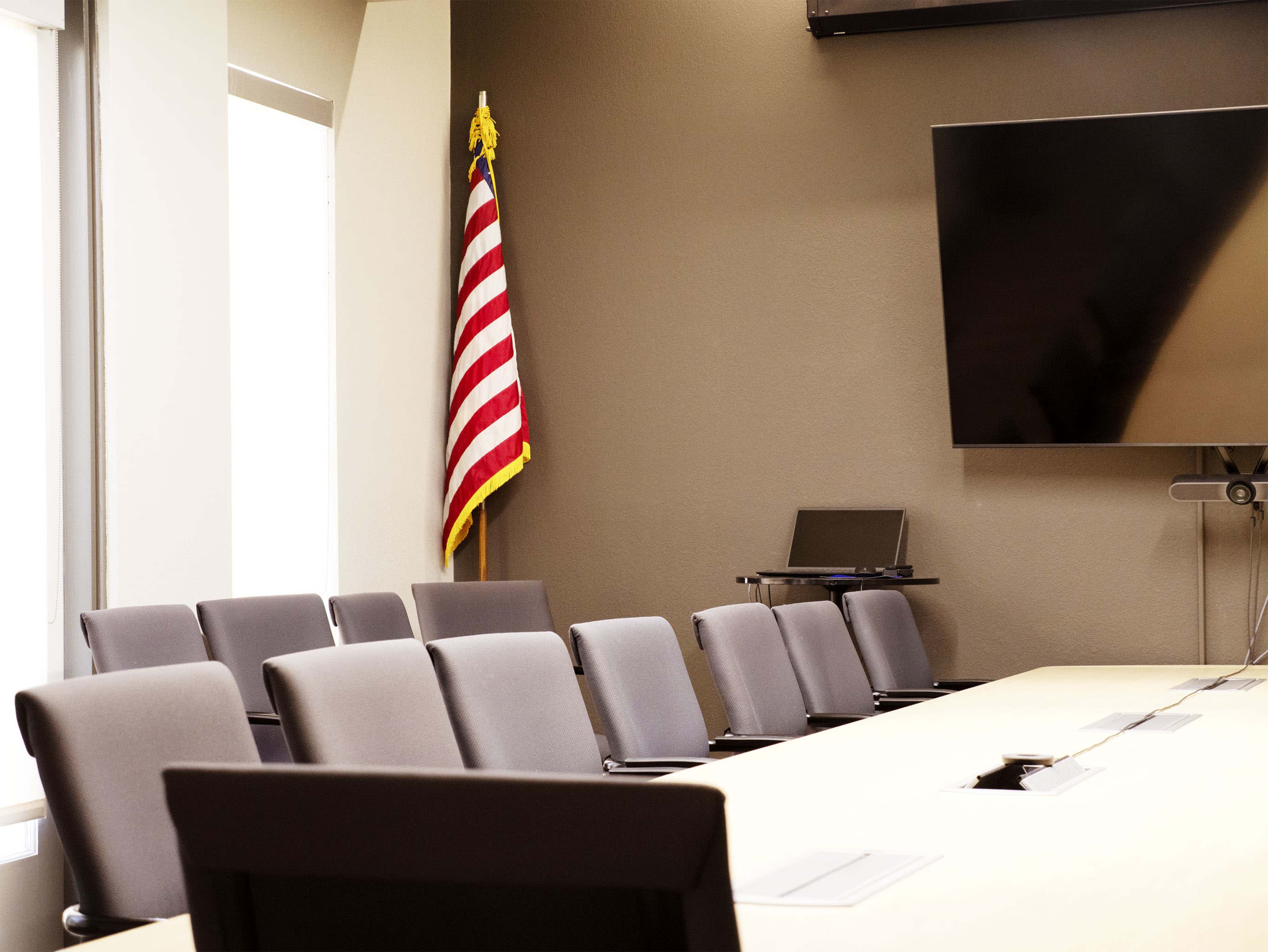 The image shows a meeting room with a long table, several gray chairs, a large screen on the wall, and an American flag in the corner.