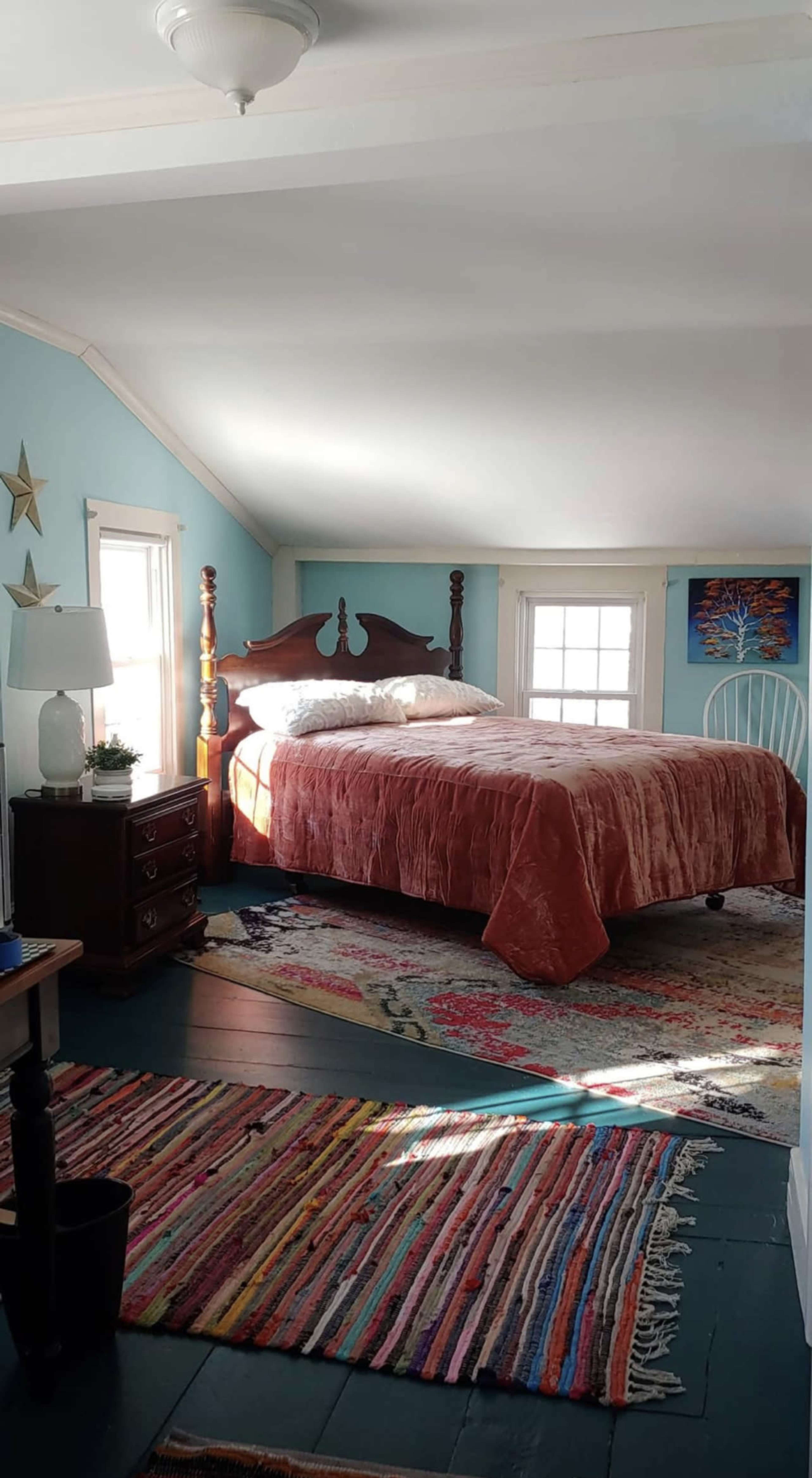The room features a bed with a wooden headboard, a colorful rug, and windows allowing natural light to enter.